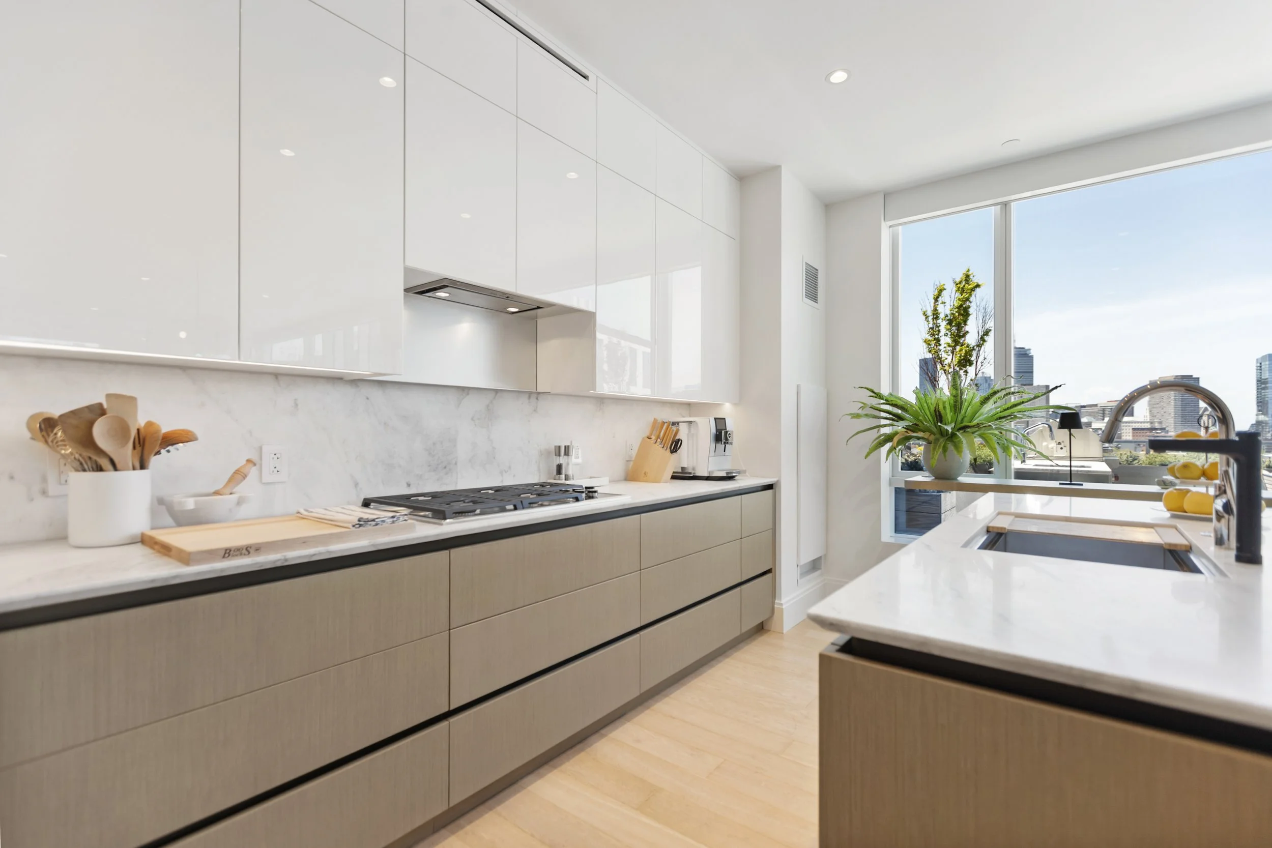Modern kitchen with white cabinets, marble countertops, a large window with city skyline view, and green plants.