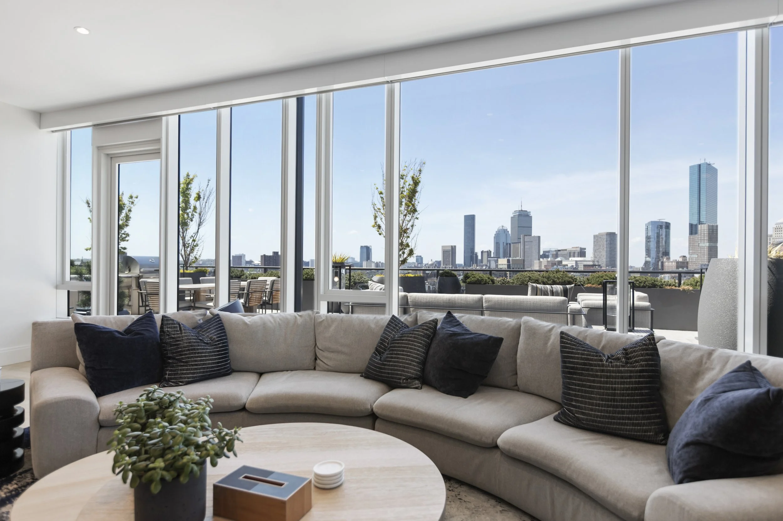 Living room with large floor-to-ceiling windows showing city skyline and blue sky, beige sectional sofa with black and blue pillows, light wood coffee table with potted succulent, sitting area with outdoor furniture on balcony.