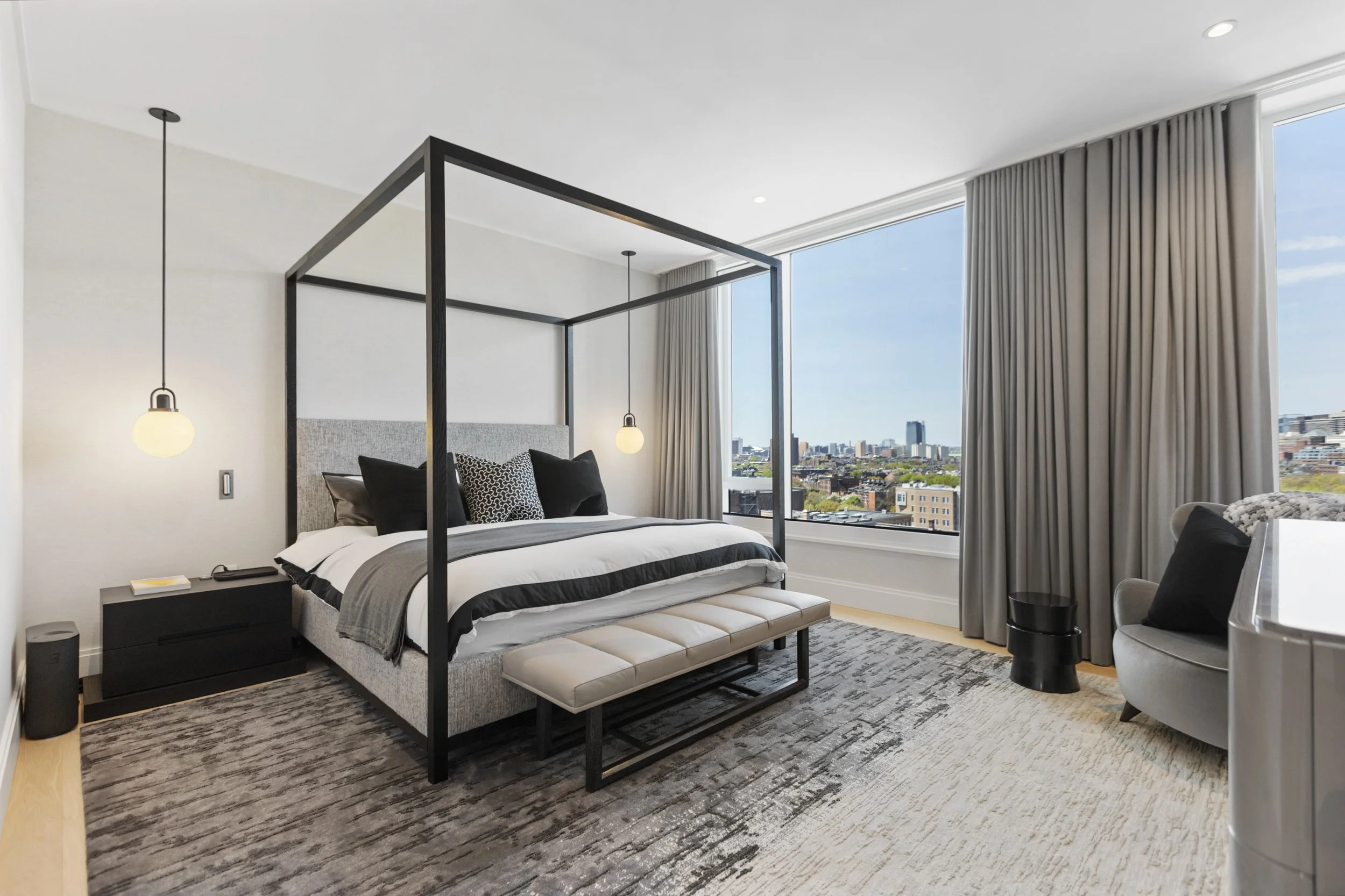 Modern bedroom with large windows, city view, black metal four-poster bed, grey upholstered headboard, black and white pillows, pendant lights, beige furniture, neutral decor, patterned rug.