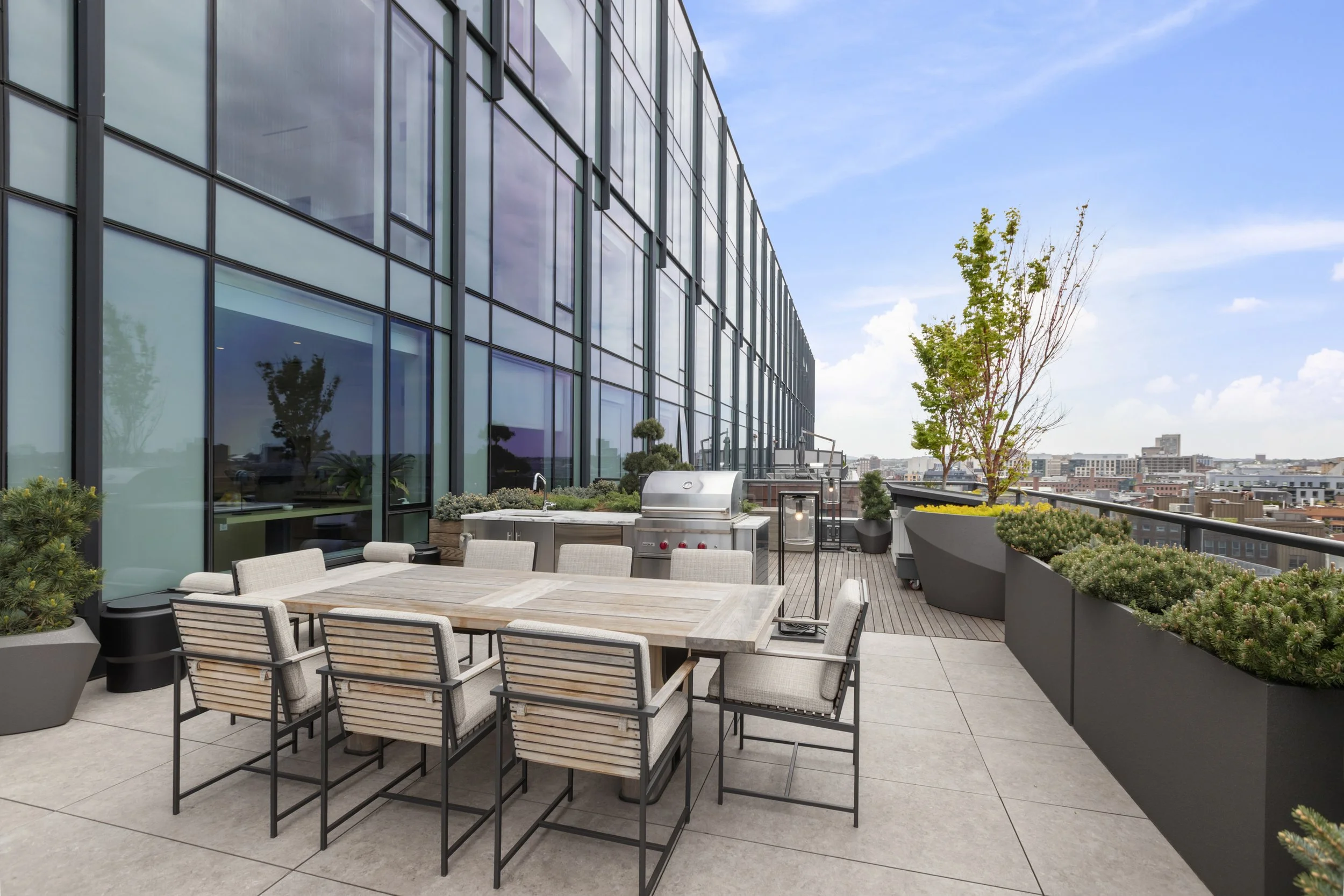 Rooftop patio with a dining table and chairs, BBQ grill, potted plants, and city skyline view with large glass building and trees.