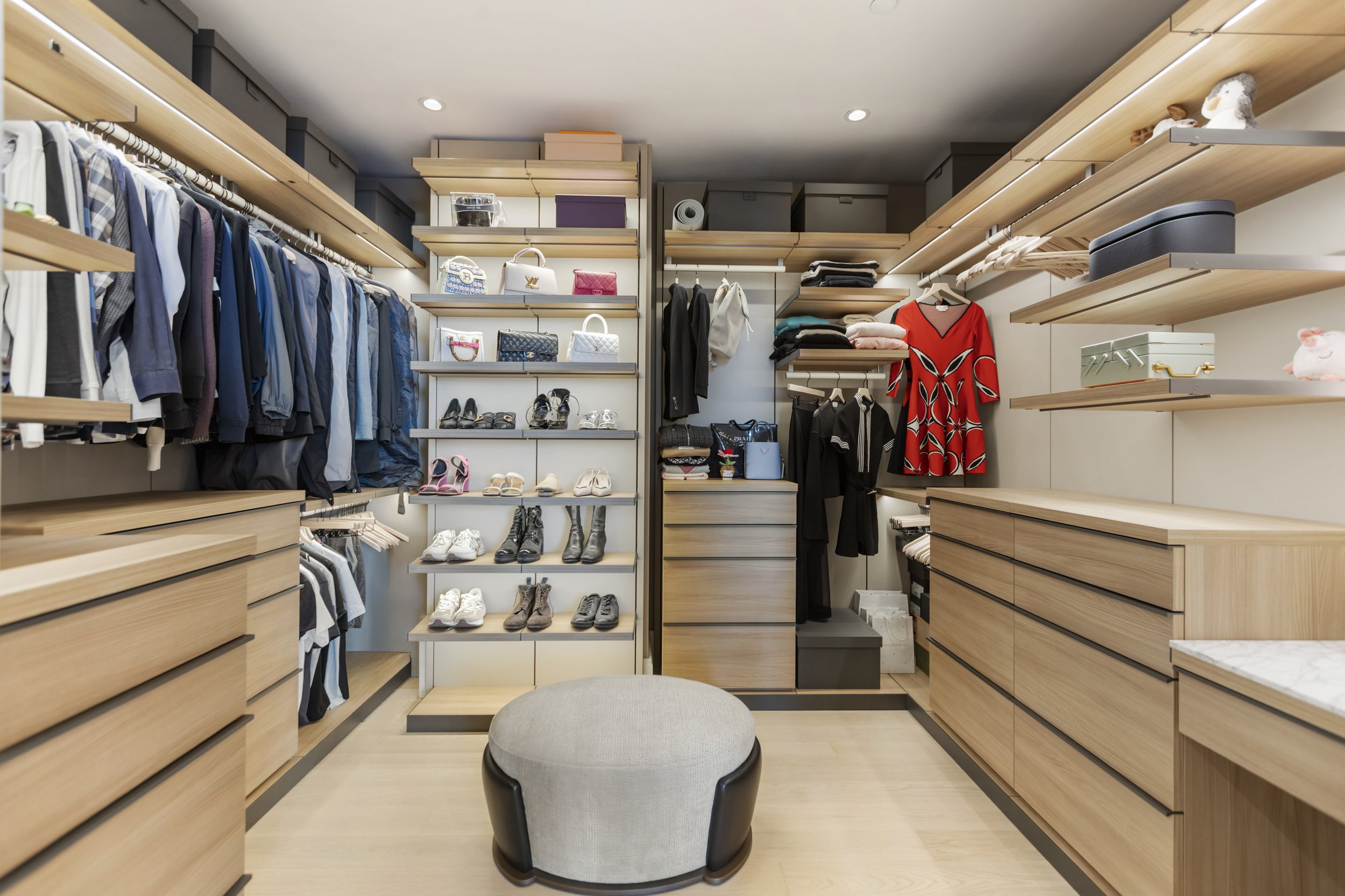 Interior of a walk-in closet with wooden shelves filled with clothing, shoes, handbags, and decorative items, a small ottoman in the center, and soft lighting.