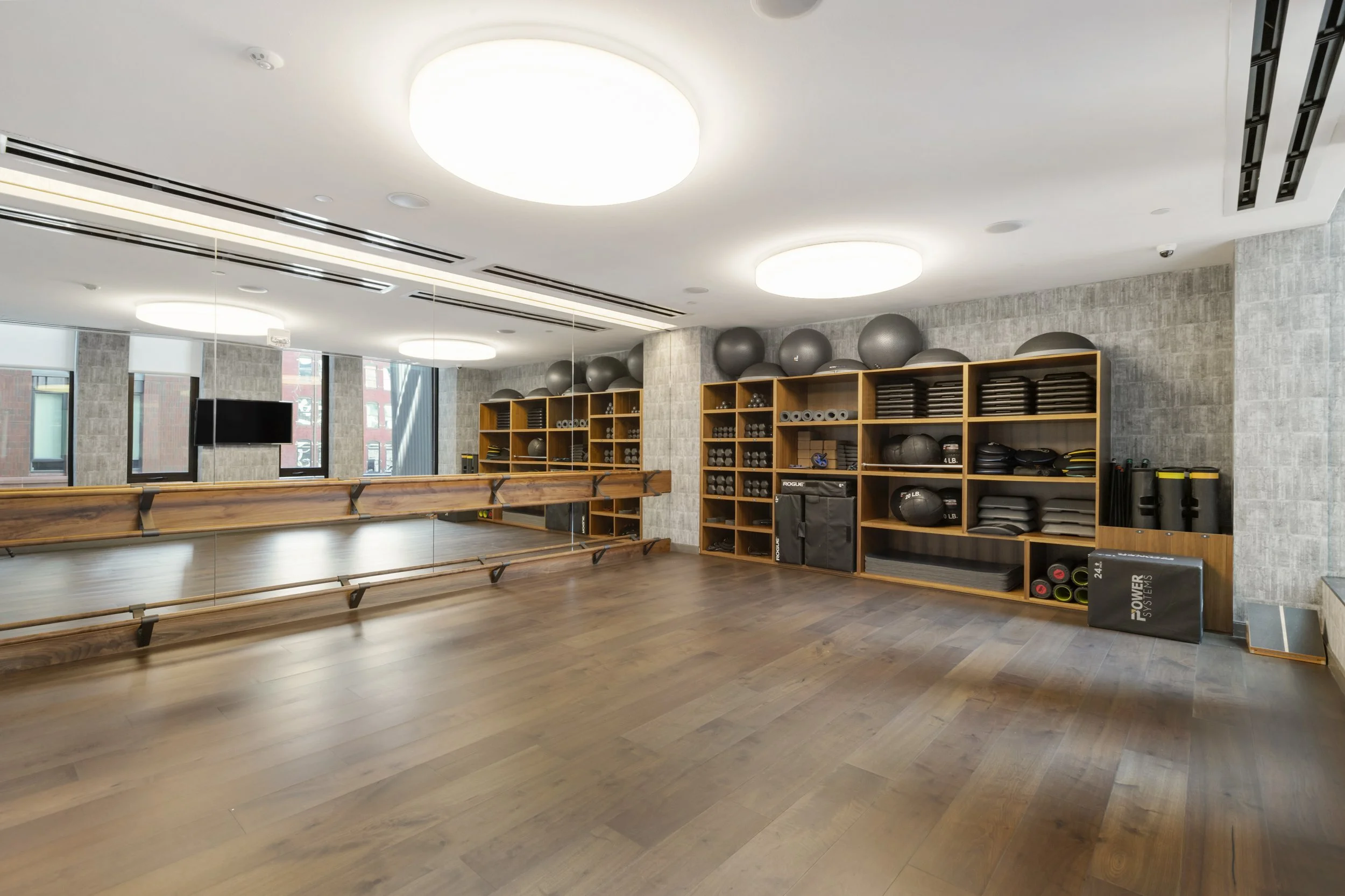 A spacious fitness studio with wooden floors, mirrored walls, ceiling lights, and shelves holding exercise balls, mats, and weights.
