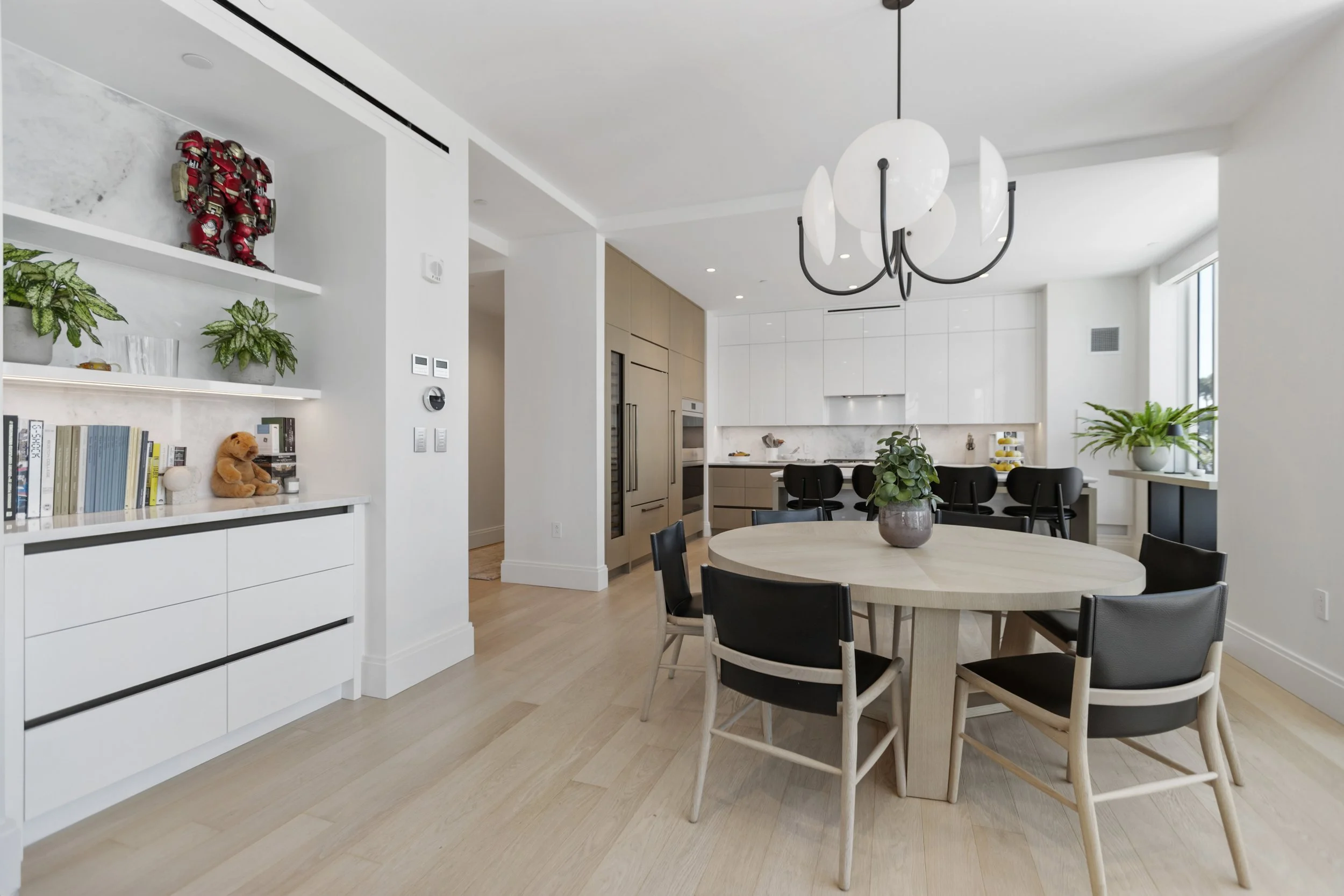 Modern open-plan kitchen and dining area with white cabinetry, a round dining table with black chairs, a large window with plants, and a contemporary chandelier.