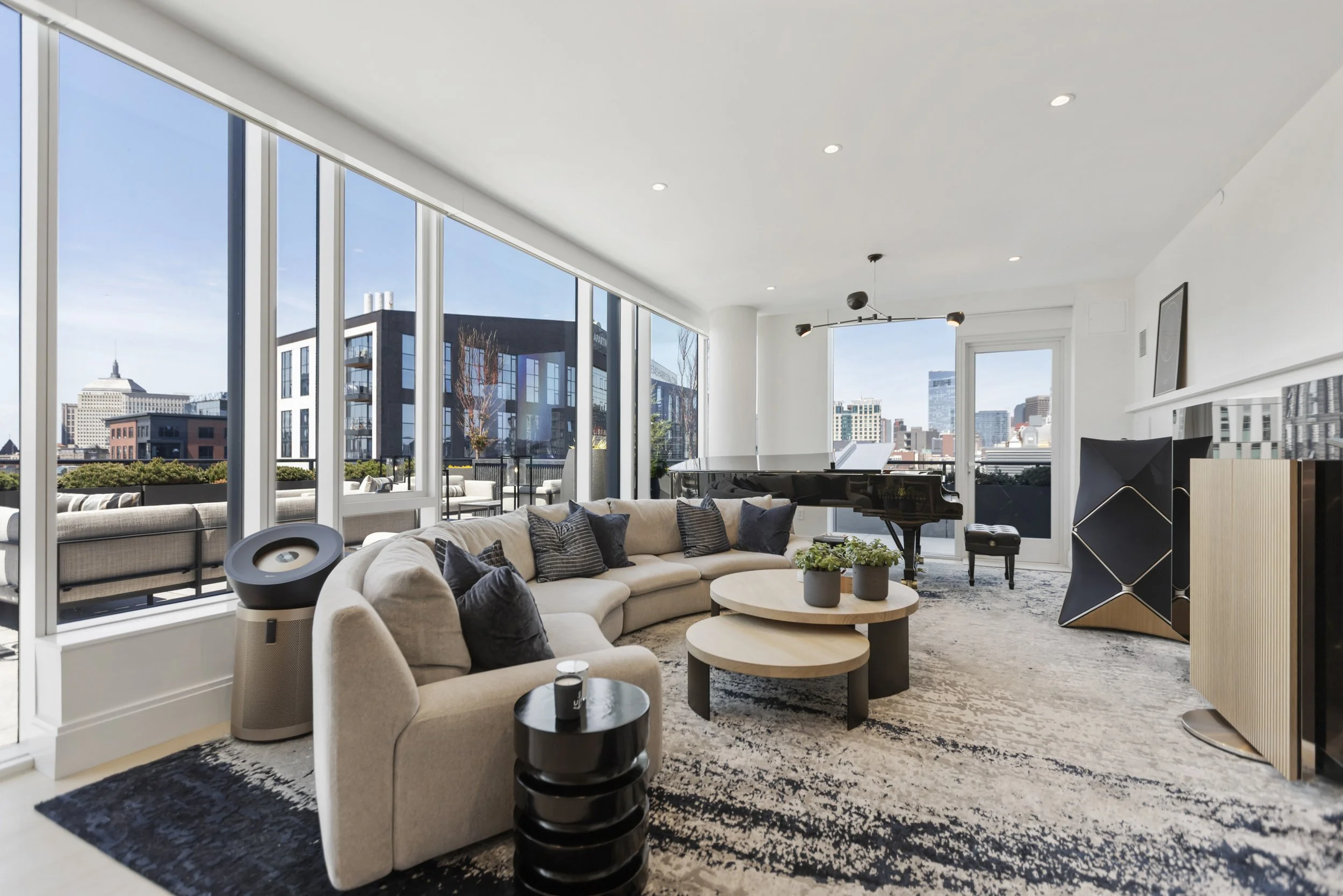 Modern living room with large floor-to-ceiling windows showing cityscape views, beige sofa with multiple cushions, black and beige abstract area rug, black side tables, and minimalistic black and wood shelving.
