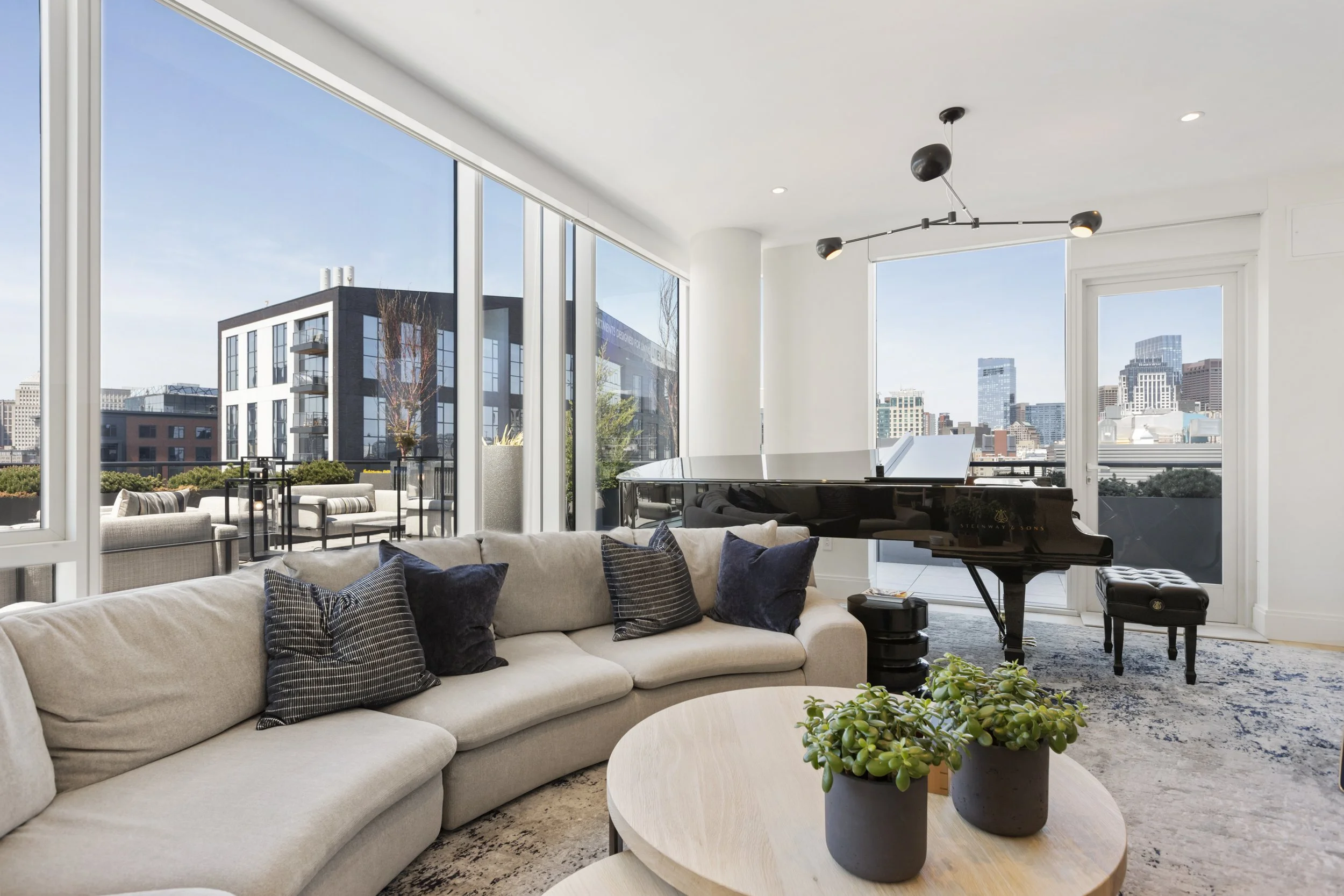 Modern living room with large windows showing city view, beige sofa with dark pillows, a black grand piano, and a wooden coffee table with potted plants.