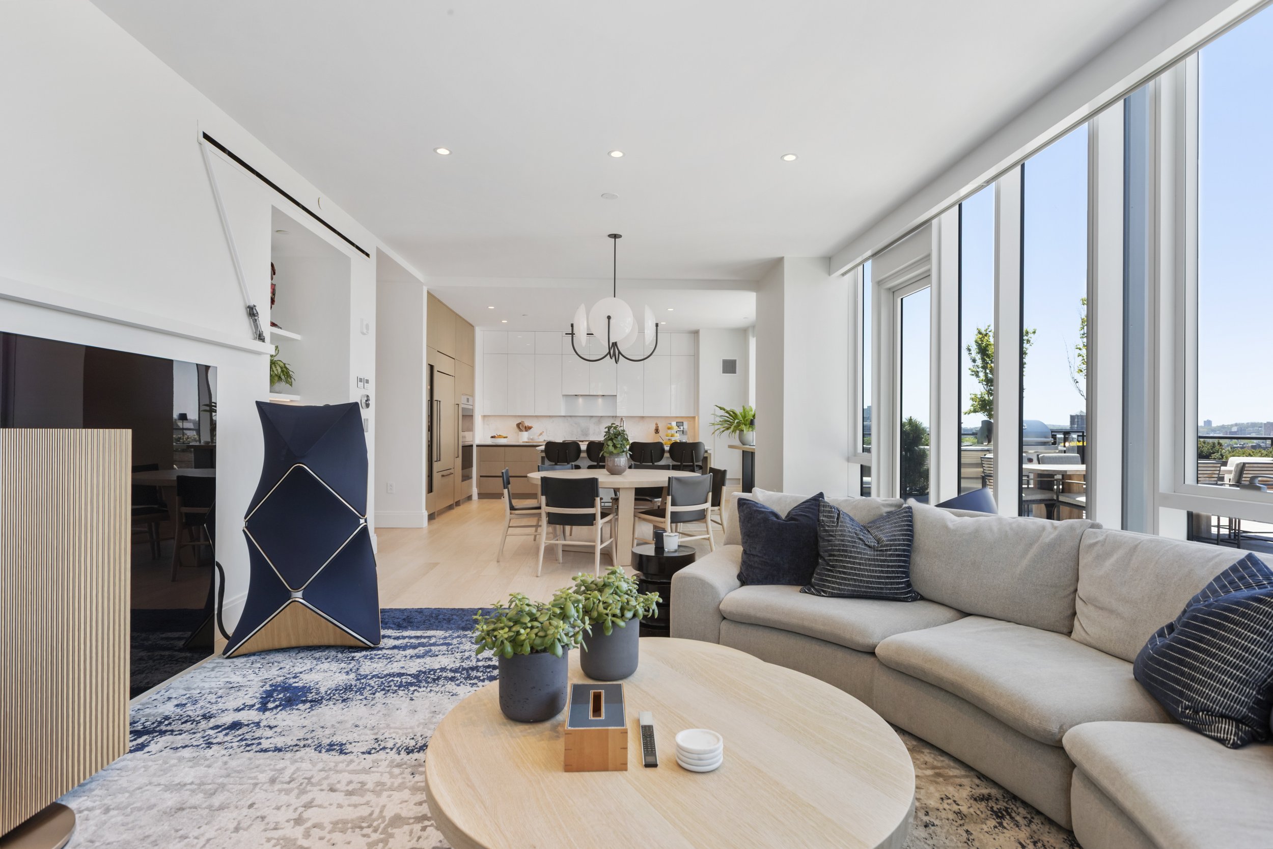 Modern open-concept living room and kitchen with large windows, white walls, wooden floors, and contemporary furniture.