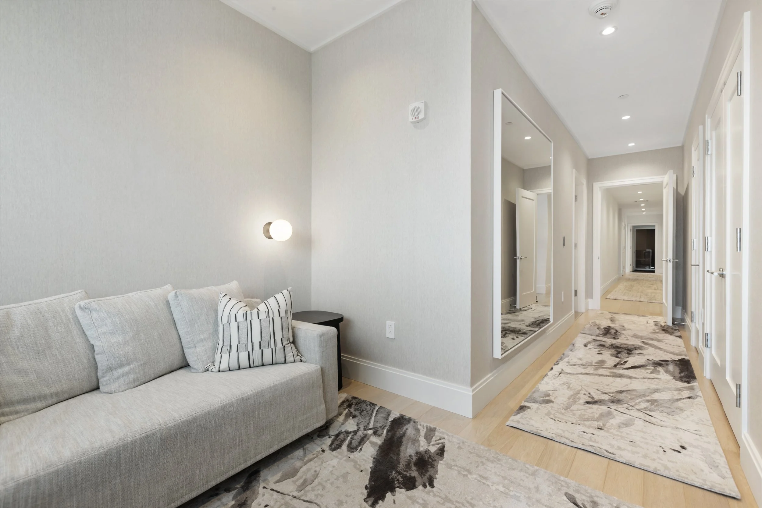 A modern living room with a light-colored sofa, striped pillows, a small black side table, and a white wall with a round wall sconce. The hallway has wooden floors, white walls, and multiple doors leading to other rooms.