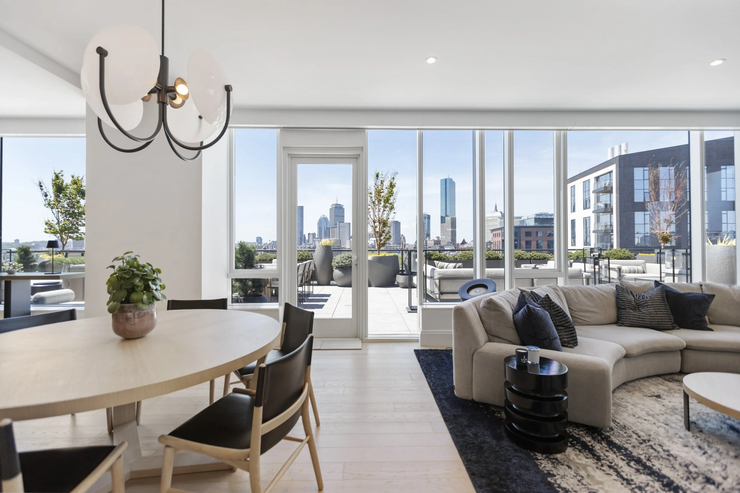 Modern living room with large windows viewing a city skyline, beige sofa with pillows, black side table, dined with a light wood table and black chairs, ceiling light fixture, plants on patio.