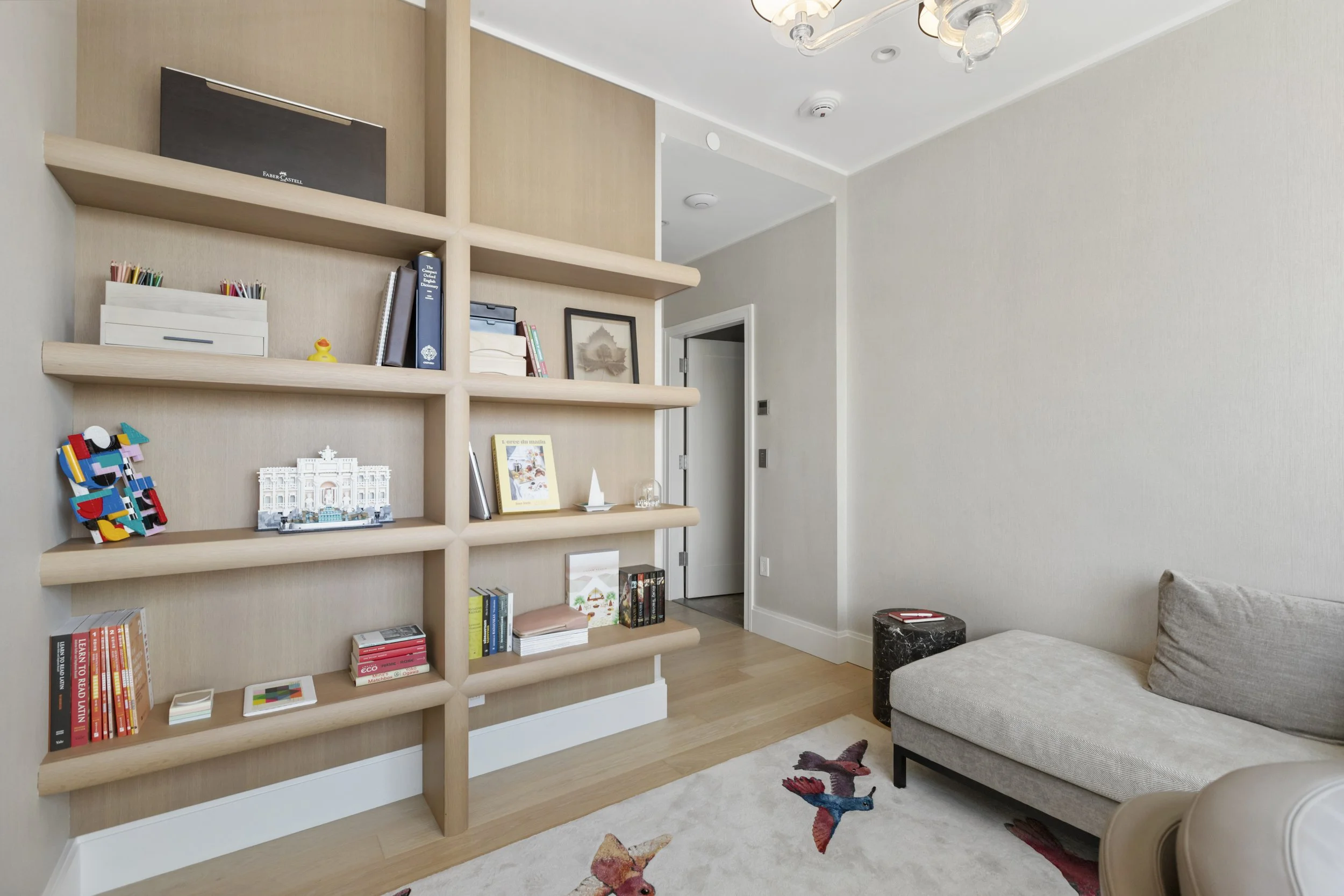 Living room with a wooden shelving unit filled with books, decorative items, and framed pictures, a gray sofa with cushions, a small black side table, and a colorful butterfly-patterned rug on the floor.