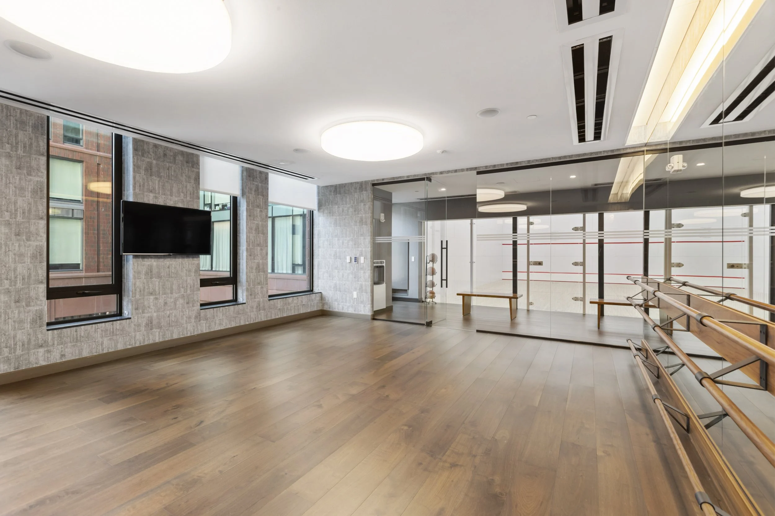 A modern dance studio with wooden flooring, ballet barres, large windows, and ceiling lights.