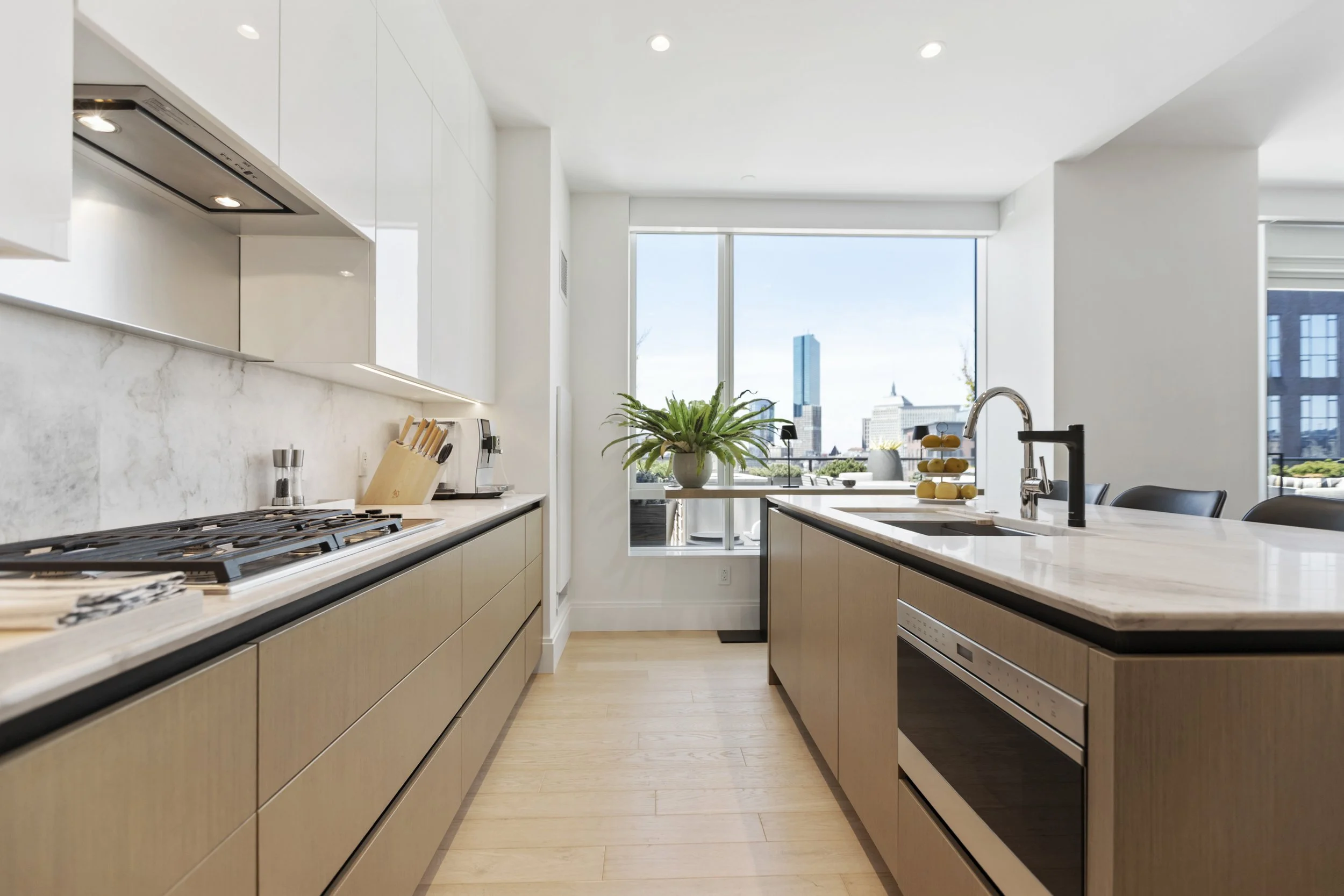 A modern kitchen with light wood cabinets, white marble countertops, a large window with a city view, and stainless steel appliances.