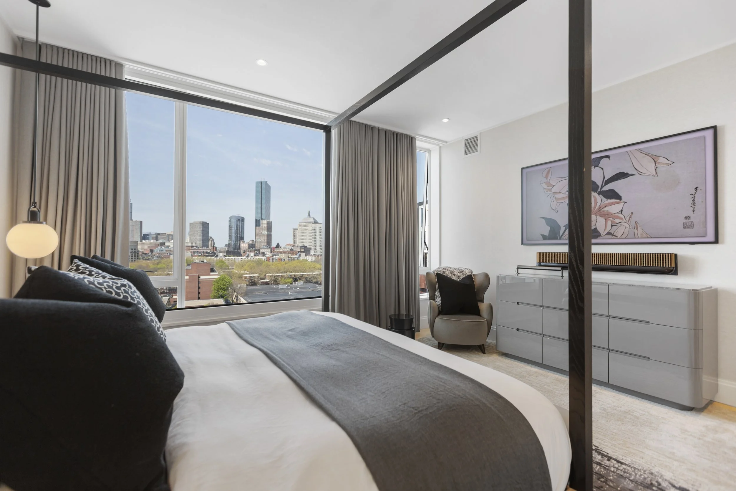 A modern hotel room with a large window showing a city skyline, a bed with black and white pillows, a nightstand, a floor lamp, and contemporary wall art.