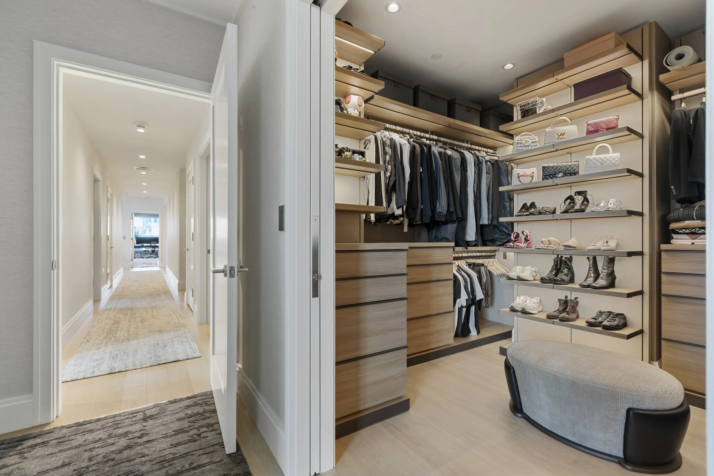A walk-in closet with clothing, shoes, and handbags on shelves and hanging rods, next to a hallway with beige carpet and white walls.