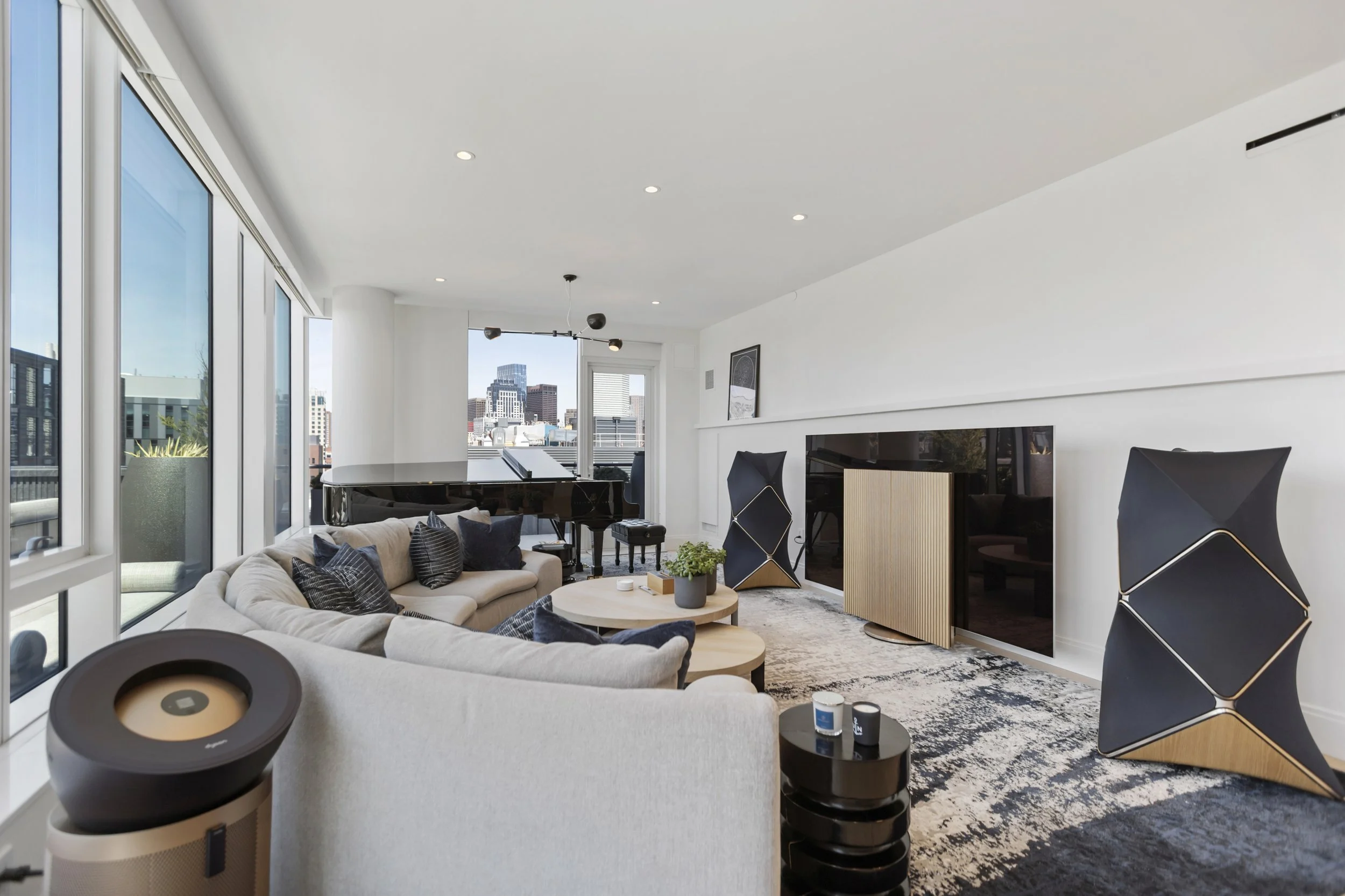 Modern living room with large windows showing city skyline, beige sectional sofa with pillows, black grand piano, black armchairs, round coffee table with plant, television, abstract artwork, and a black and wood fireplace.