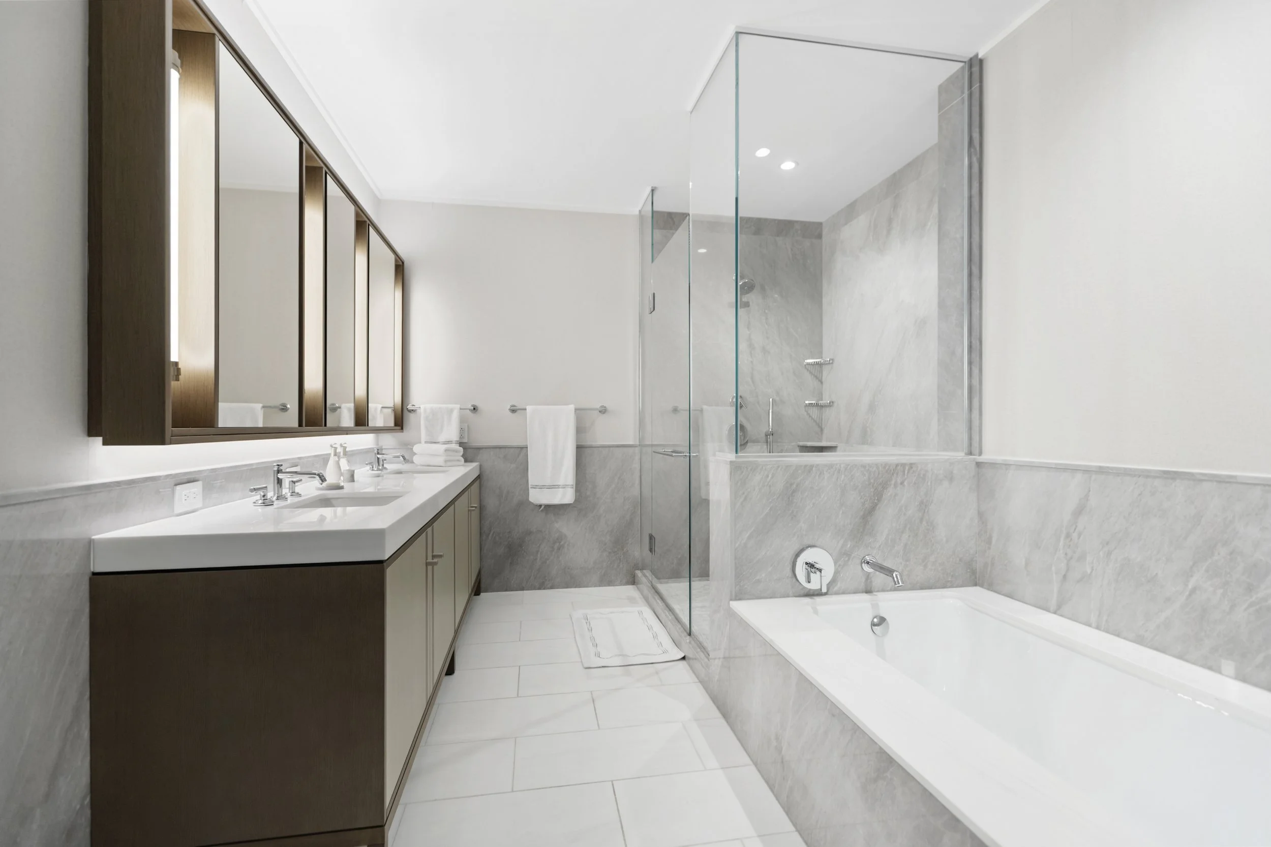 Modern bathroom with double vanity, wooden cabinets, large mirror, glass-enclosed shower, and bathtub with marble walls.