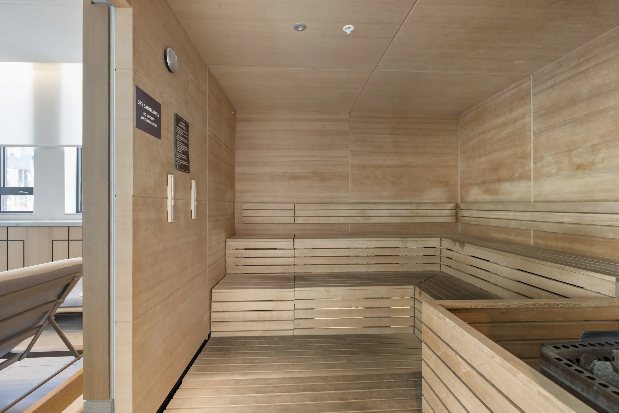 An empty modern sauna room with wooden benches and walls, a small heater, and signs on the wall. There is a window showing a building outside.
