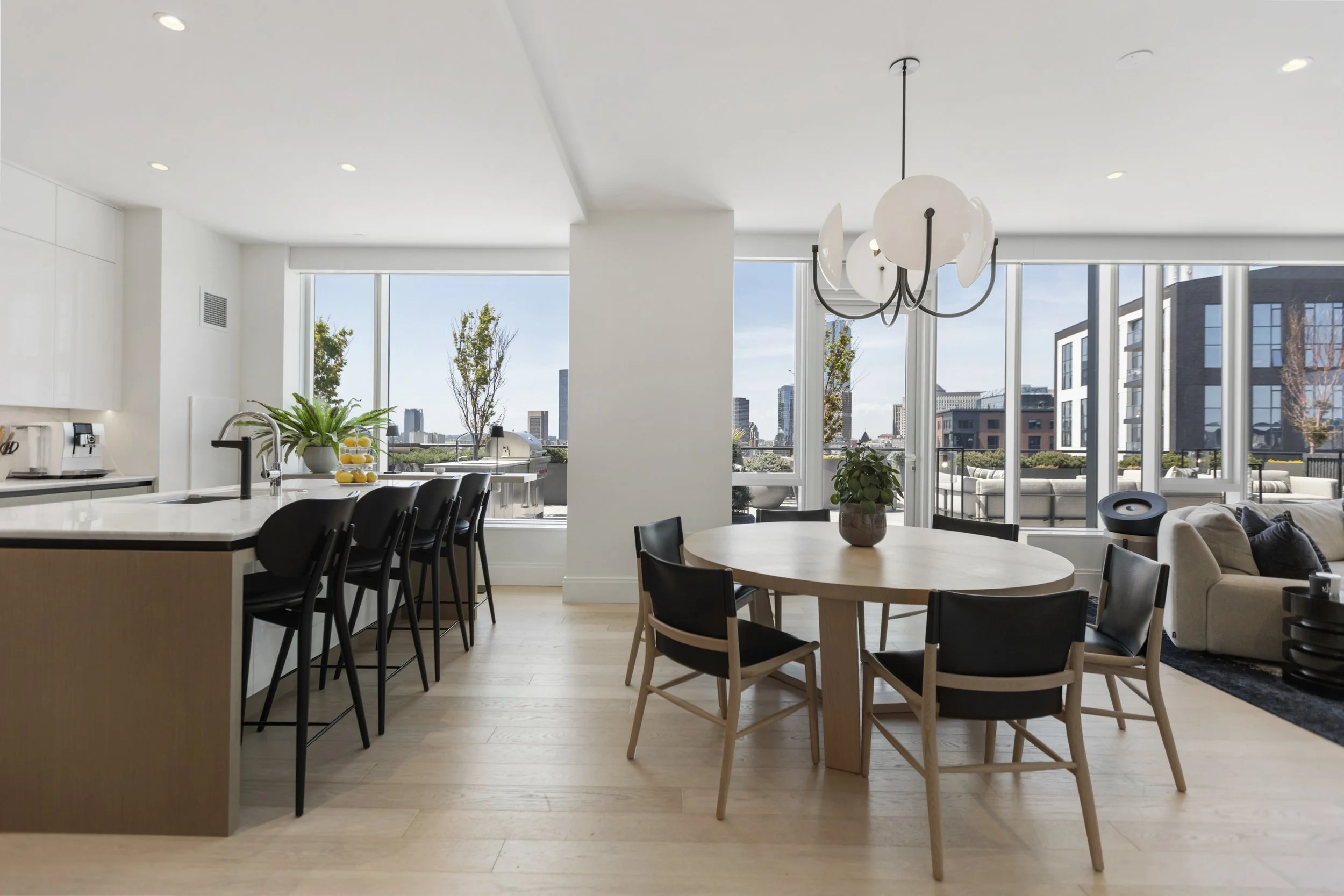 Modern open-plan living space with kitchen island, black chairs, round wooden dining table with a potted plant, large windows with cityscape view, hanging pendant light, and beige sofa.