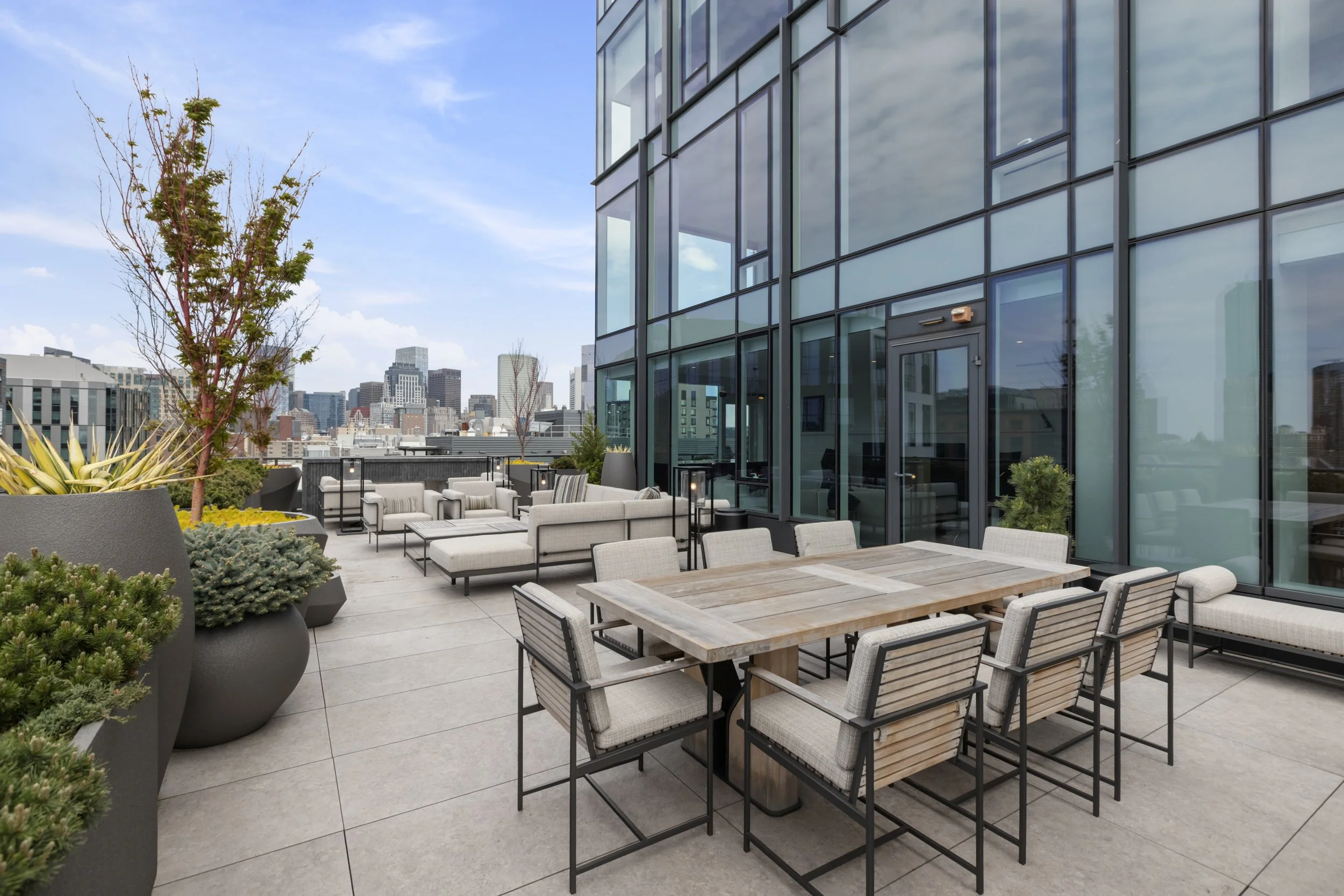 City rooftop outdoor terrace with modern seating area, large planters with greenery, and glass building exterior reflections, city skyline in the background under a blue sky with clouds.