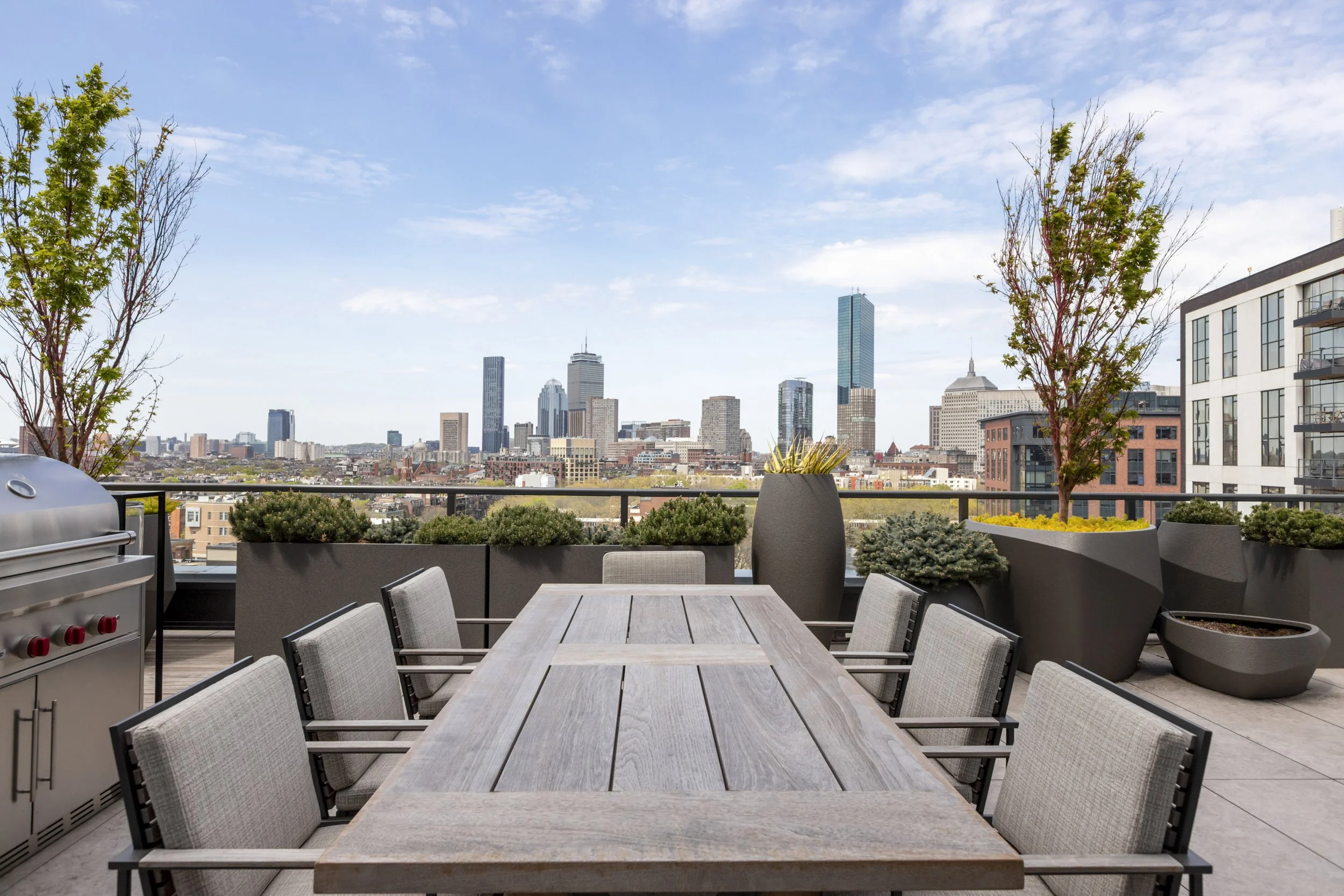Views of a city skyline with tall skyscrapers from a rooftop patio that has a long wooden dining table, gray chairs, potted plants, and a barbecue grill, under a partly cloudy sky.
