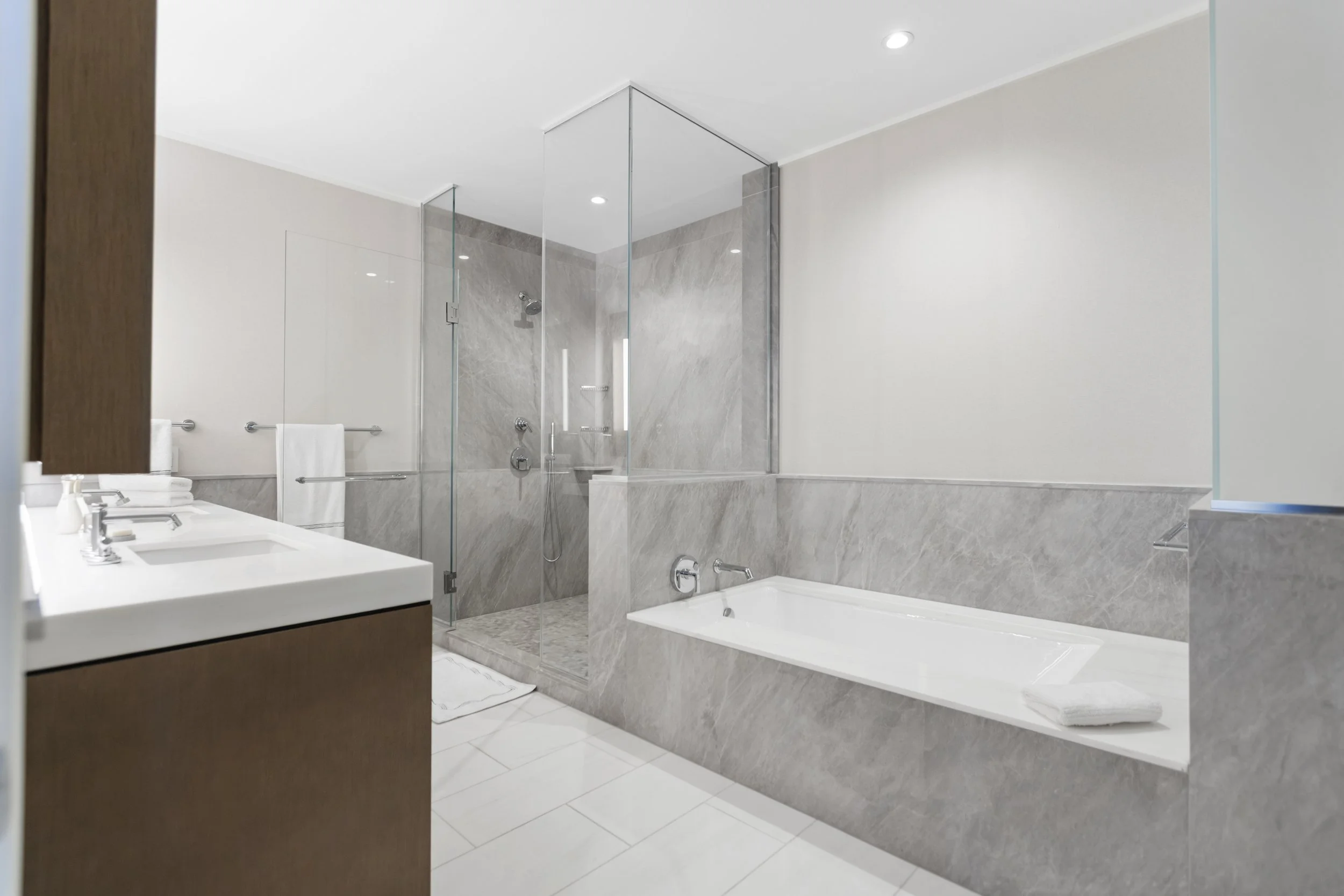 Modern bathroom featuring a built-in bathtub and a glass-enclosed shower with grey marble tiles, white ceiling with recessed lighting, a white double sink vanity, and white walls.