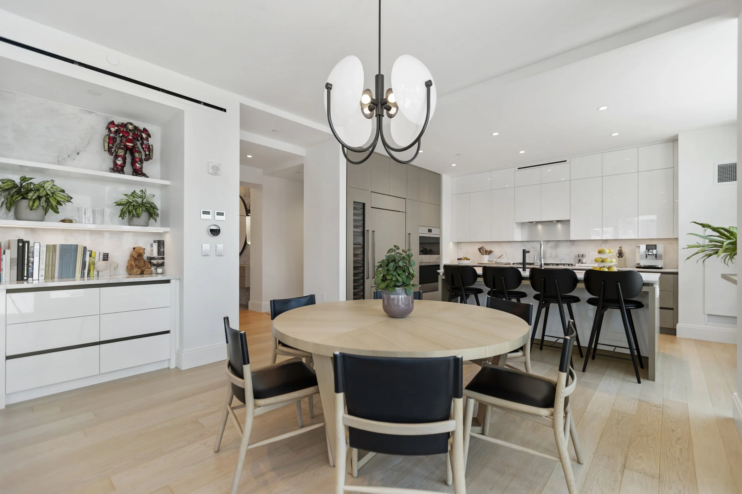 Modern kitchen and dining area with white cabinets, black chairs around a round wooden table, potted plants, and contemporary lighting. Decor includes a toy robot on a shelf and books.
