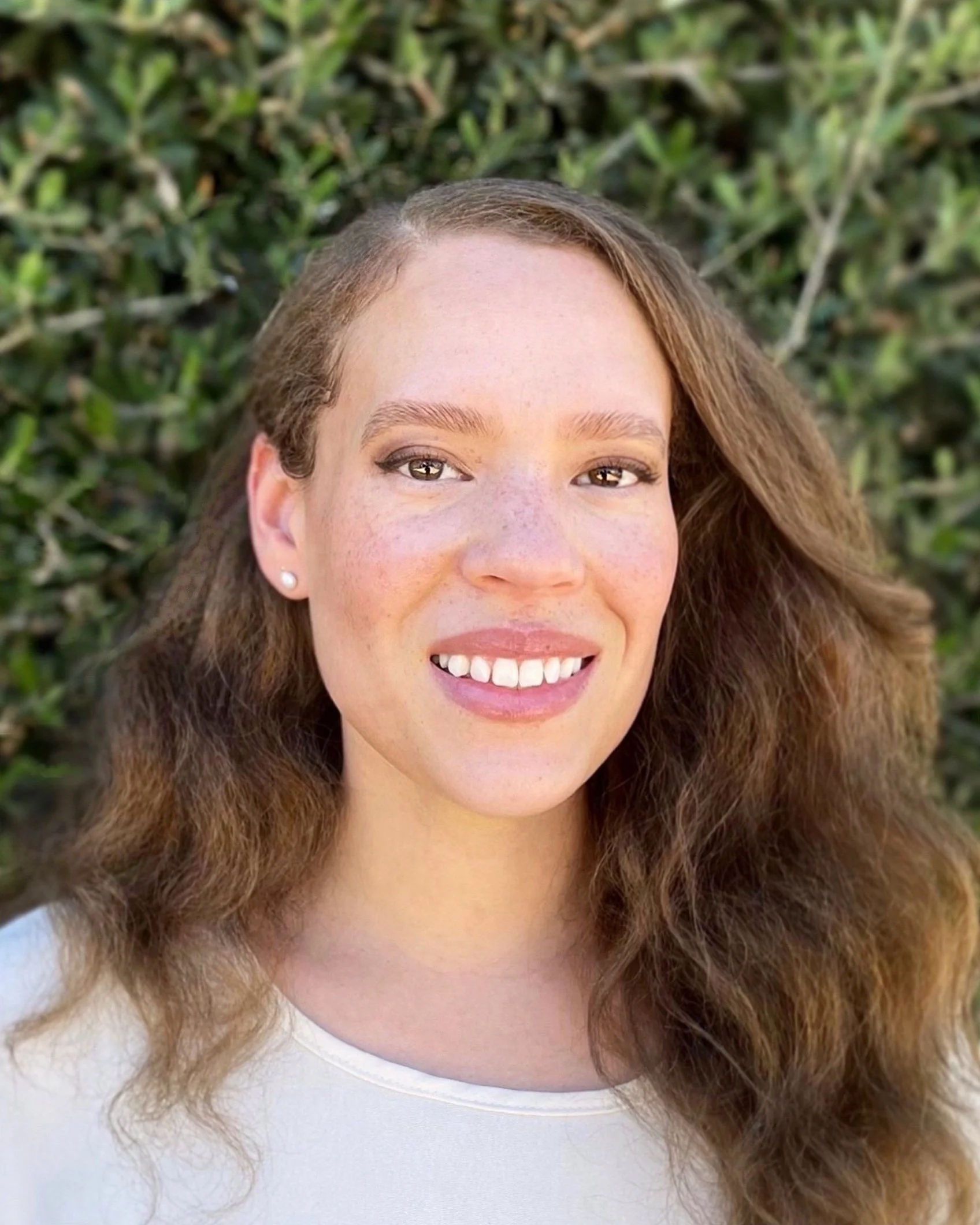 Close-up of a woman's face with brown curly hair, smiling, with greenery in the background.