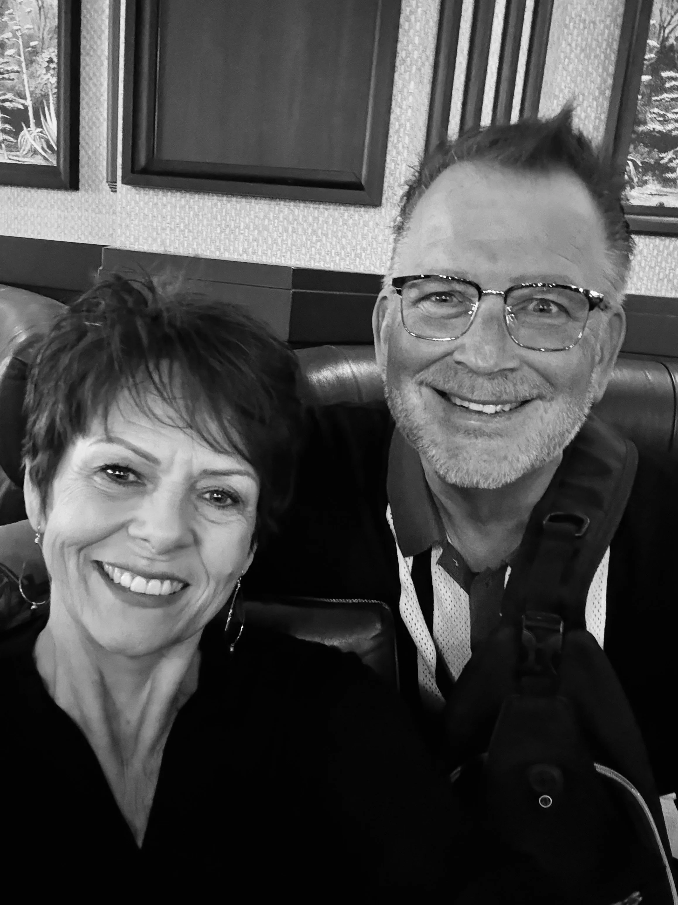A smiling man with glasses and a woman with short hair smiling. Both are in a restaurant with framed artwork on the wall behind them.
