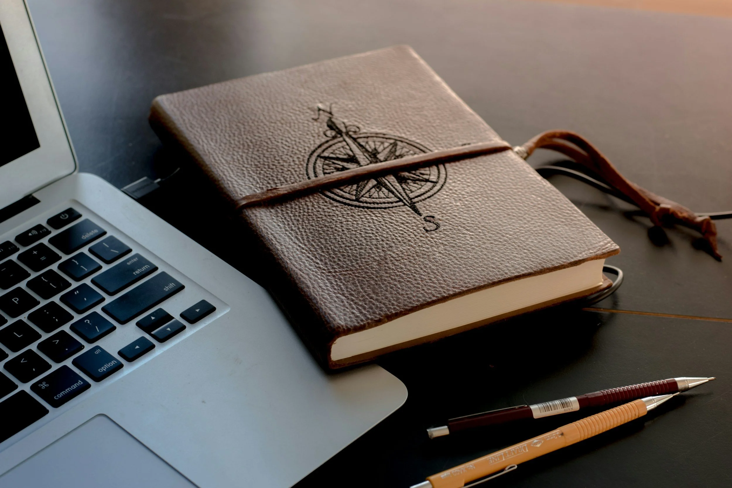 A view of a workspace featuring a silver laptop, a closed textured notebook with a compass design on the cover, and several pens on a dark surface.
