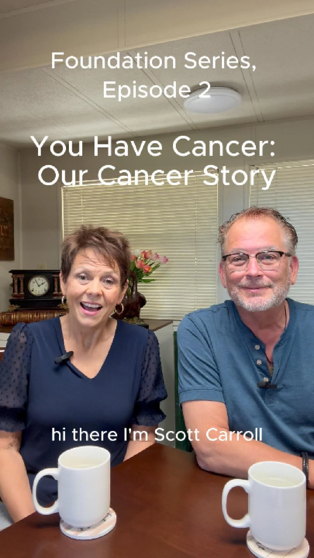 Scott & Patty Carroll sitting at a table with white mugs, smiling for a video recording. A woman with short brown hair and a navy blouse, and a man with glasses and a blue shirt are in a room with blinds and a clock on the wall in the background.