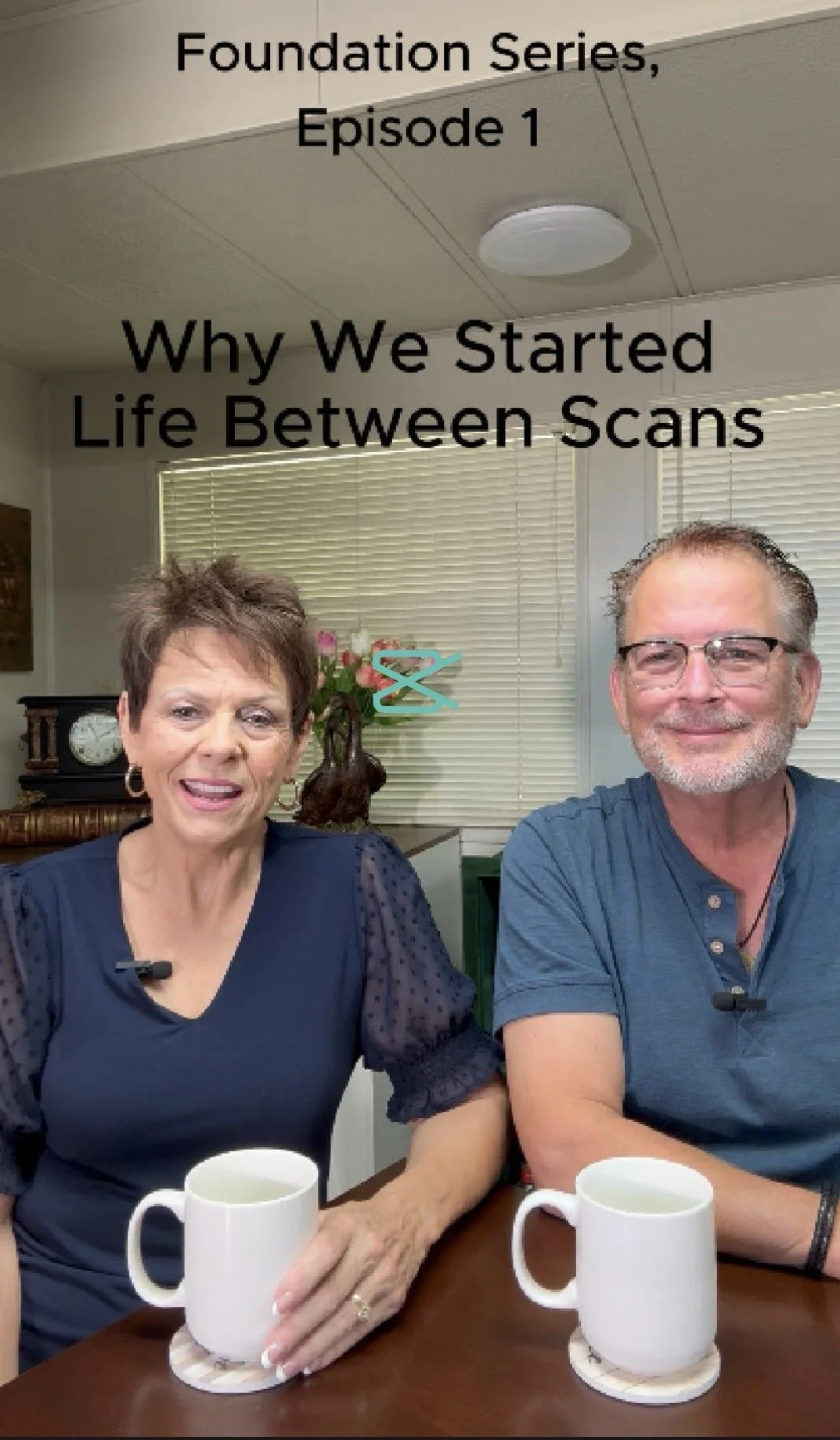 Scott & Patty Carroll sitting at a table with white mugs, smiling, in an indoor setting with blinds, a clock, and a vase with flowers in the background under a sign that reads 'Foundation Series, Episode 1 Why We Started Life Between Scans'.
