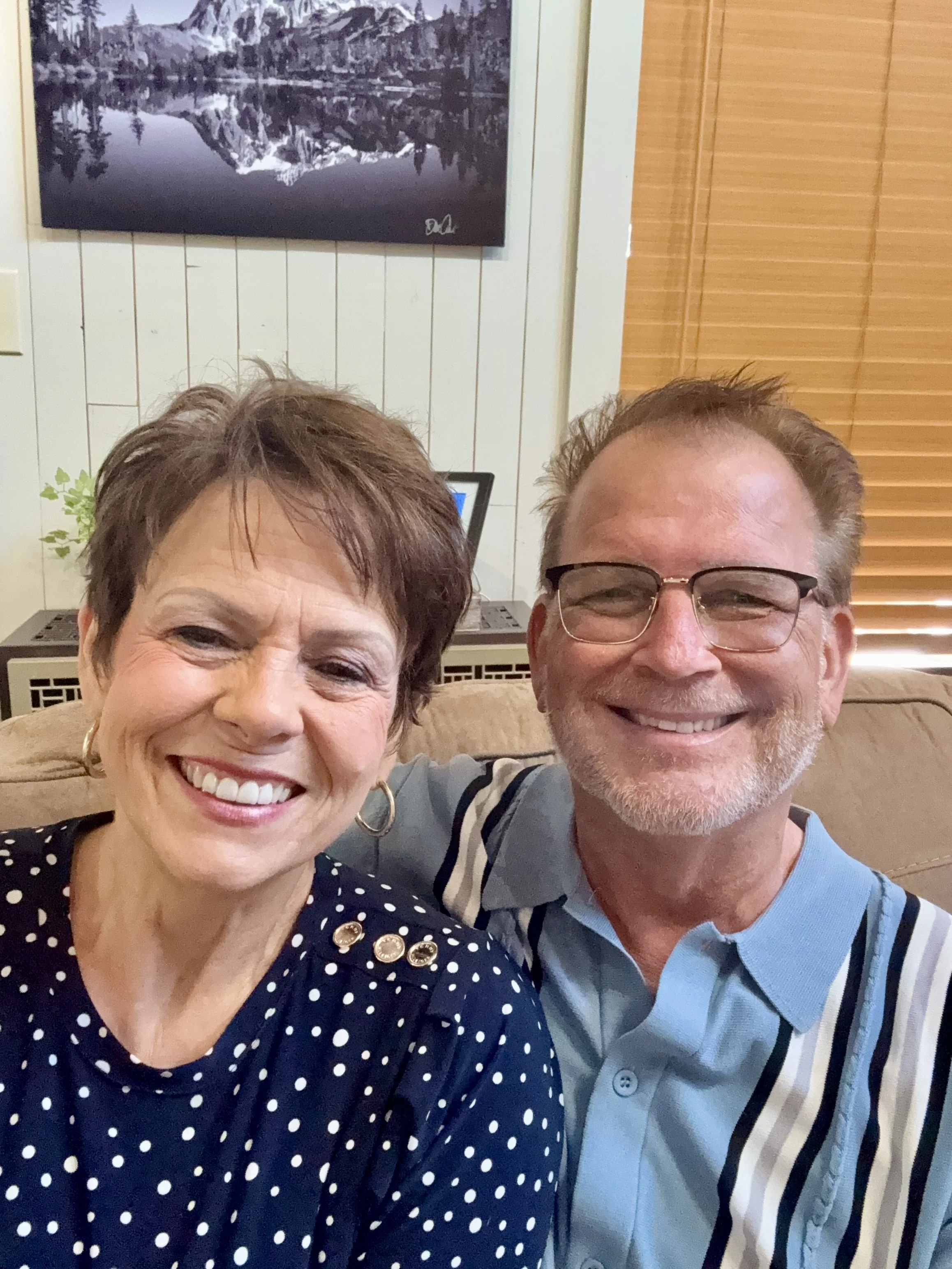 A smiling woman with short brown hair wearing a navy blue polka-dot blouse and gold earrings, and a smiling man with glasses and short light brown hair, sitting on a beige couch in a room with white paneled walls, a television, and wooden blinds in the background.