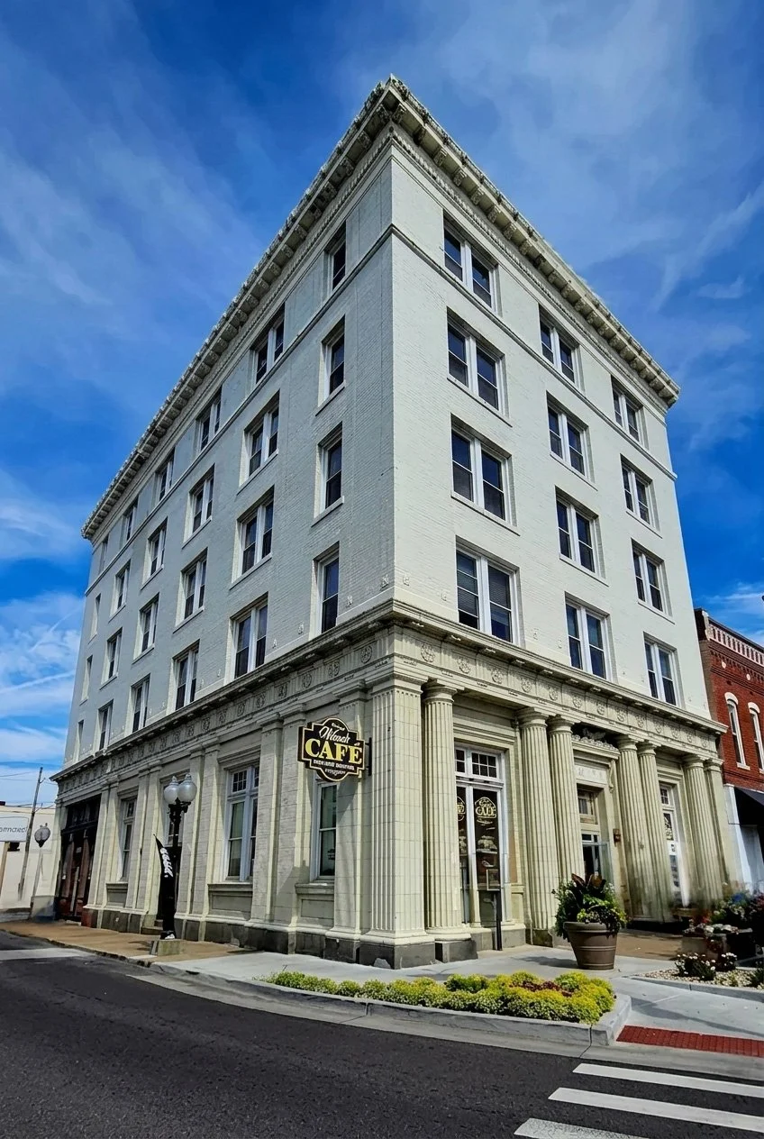 A tall, white multi-story building with classical architectural details at the corner of a street. The ground floor features a cafe with a sign reading 'Alcona Cafe,' large windows, and decorative columns. The sky above is blue with some clouds.