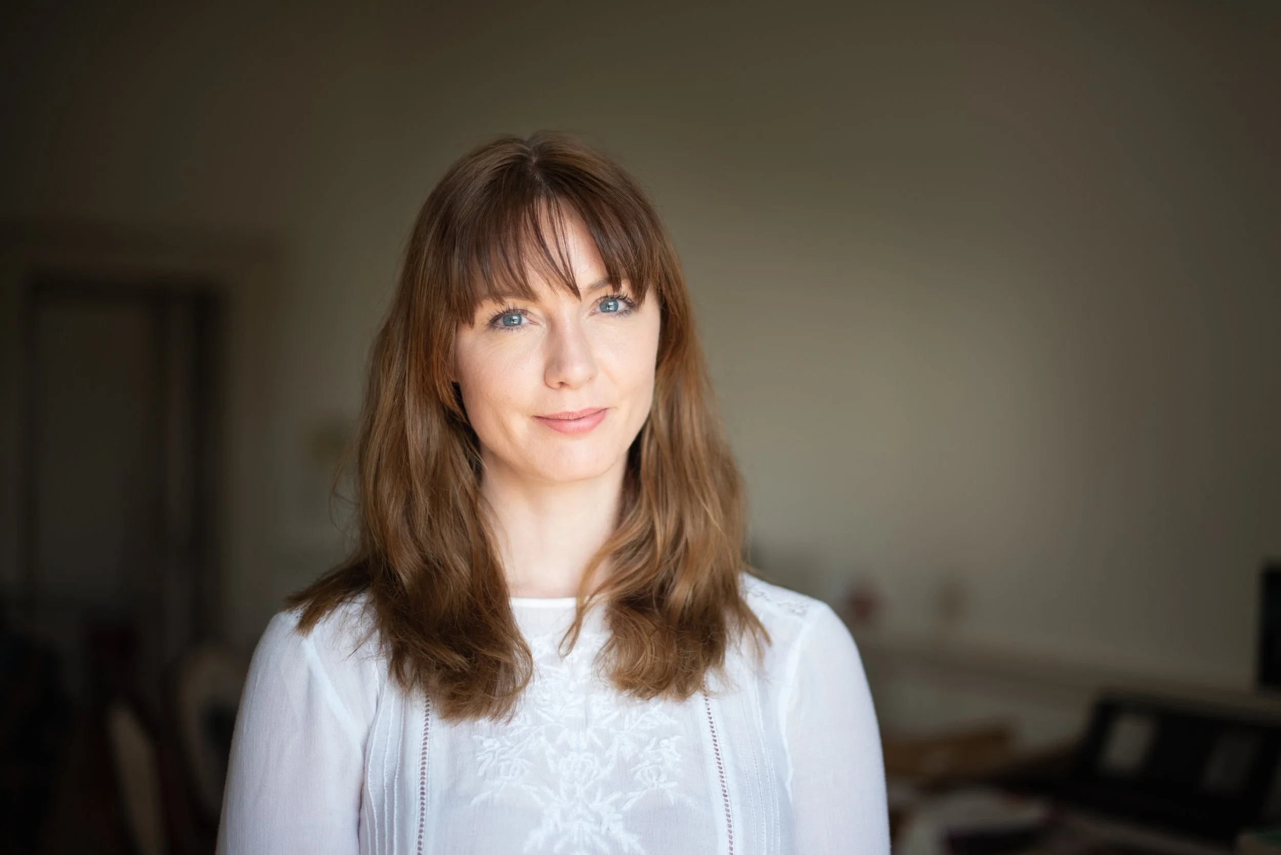 A woman with light brown hair and blue eyes wearing a white top, standing indoors with a blurred background.
