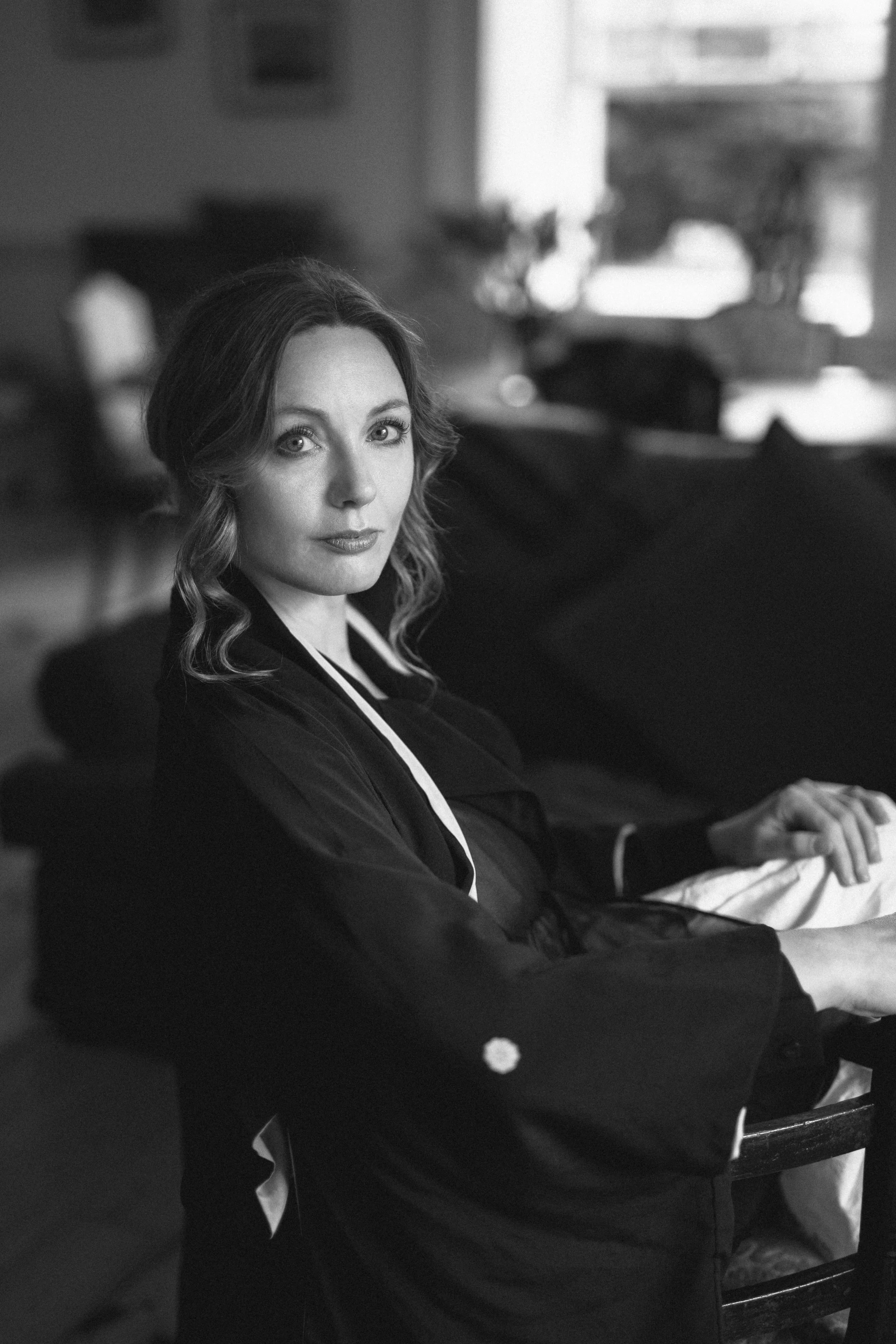 Black and white photo of a woman with wavy hair and light eye color, sitting at a table in a relaxed environment, looking directly at the camera.
