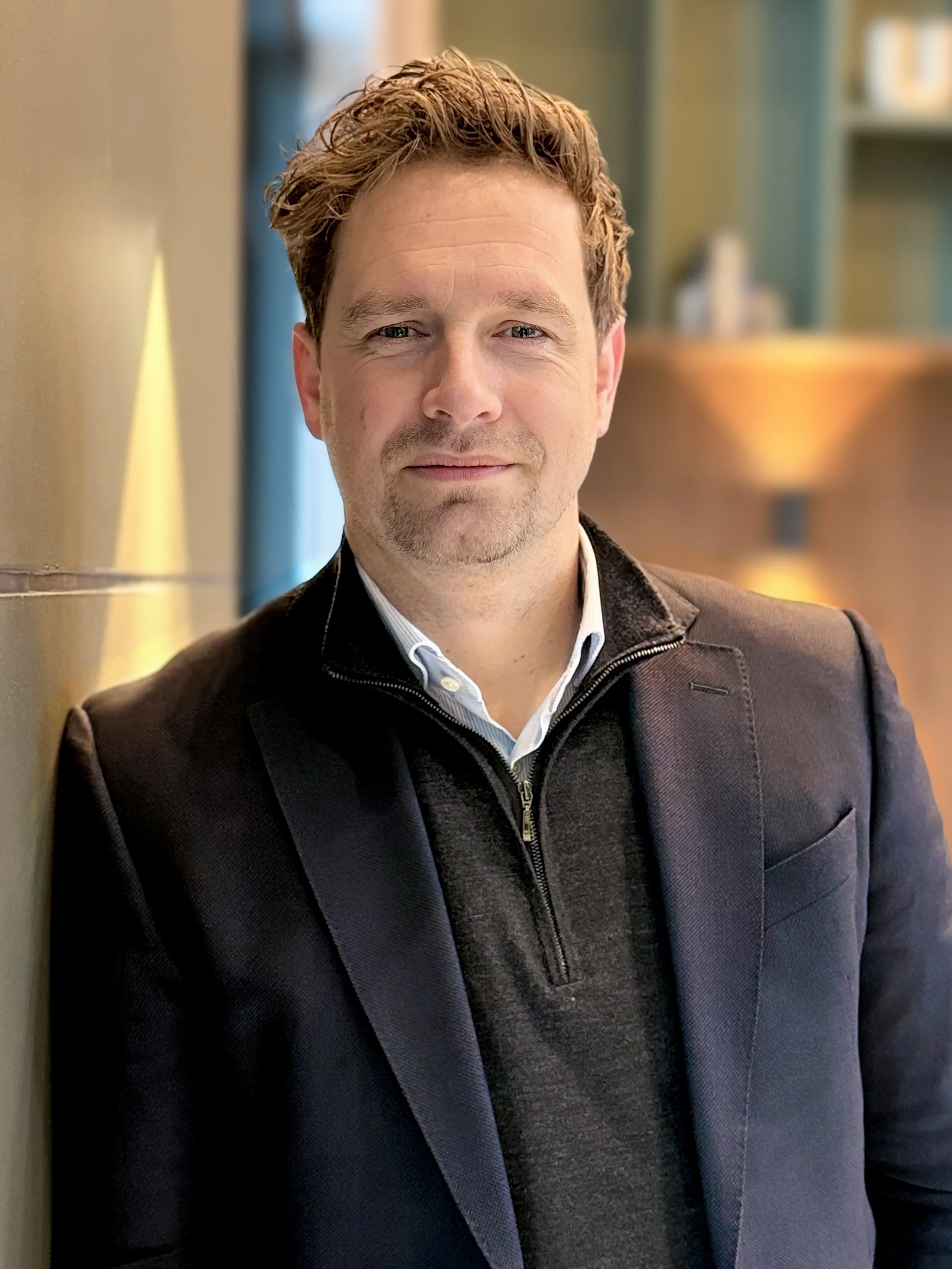 A man with short, wavy brown hair, wearing a dark jacket and a light collared shirt, standing indoors against a blurred background.