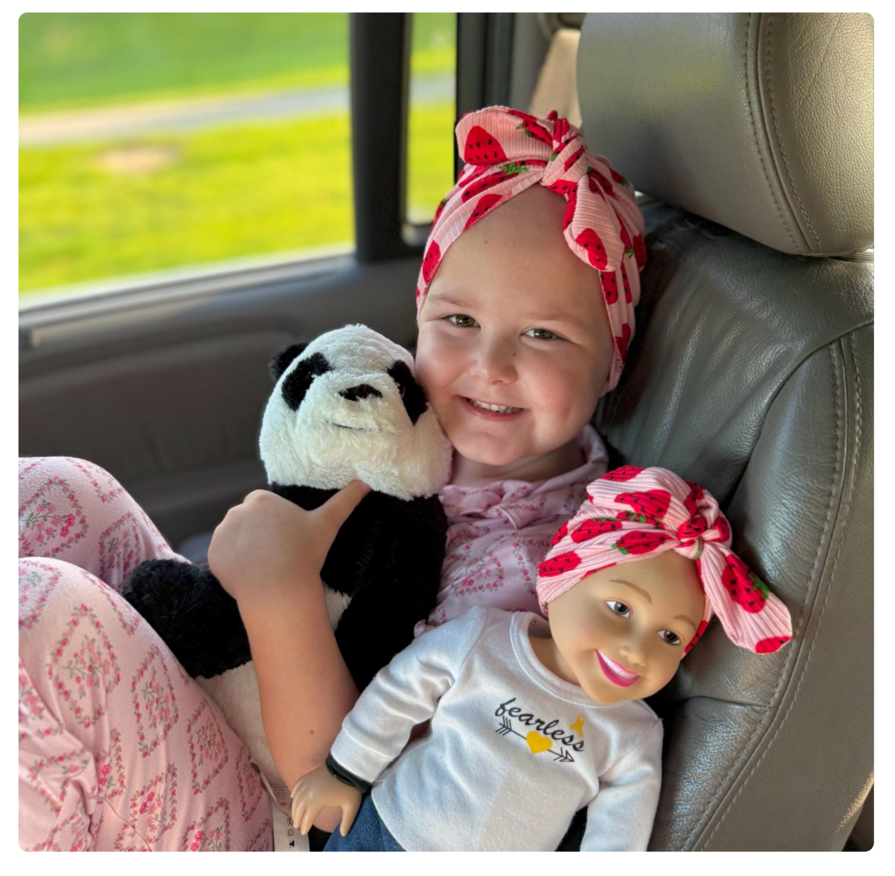 A young girl with a pink headscarf with red strawberries, smiling and holding a panda stuffed animal, sitting in a car with a green outdoor scene visible through the window.