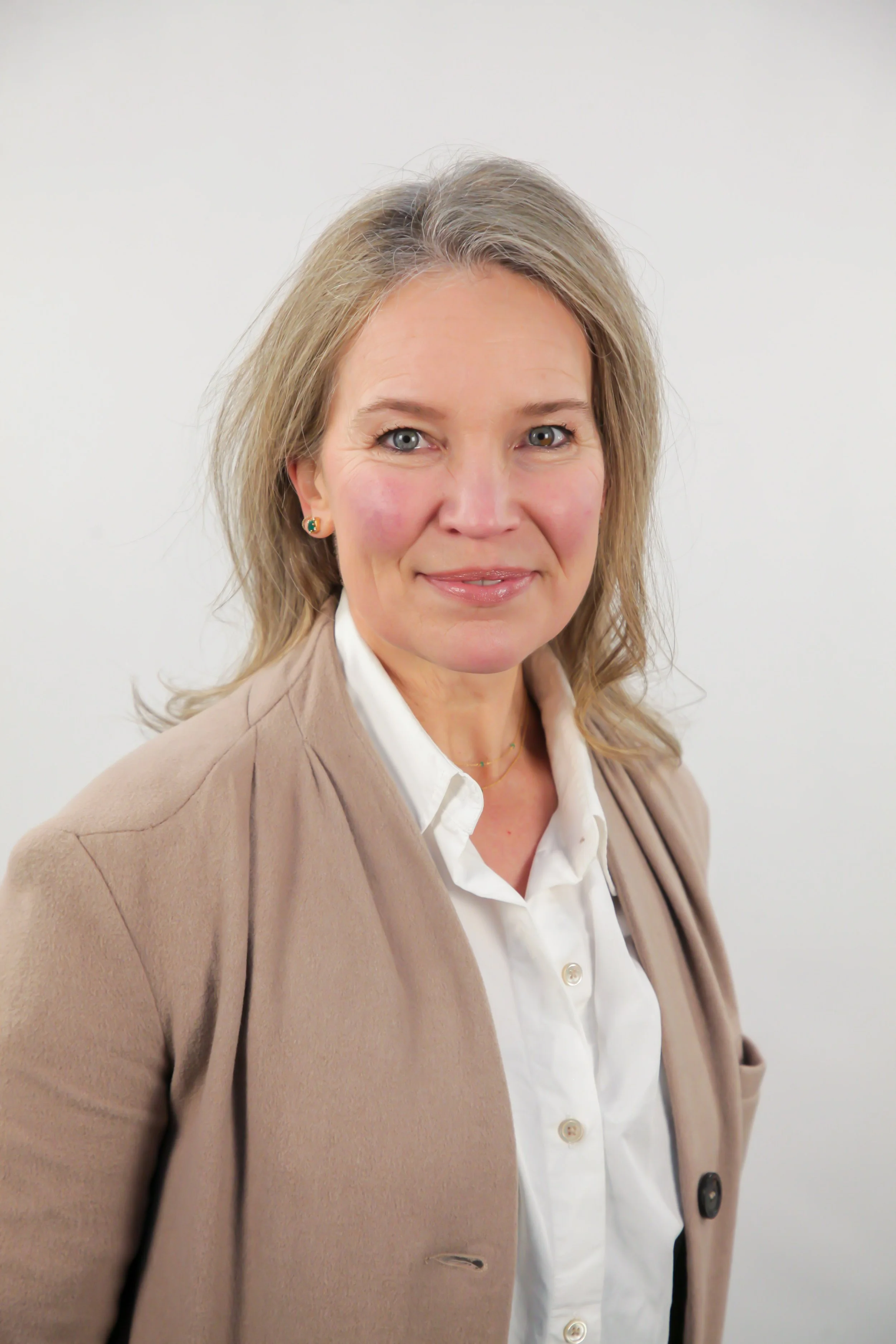 A woman with shoulder-length blonde hair, blue eyes, and earrings, wearing a beige blazer over a white shirt, standing against a plain white background.