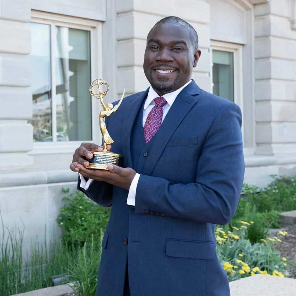 A man in a navy suit and red tie smiling while holding an Emmy award trophy outdoors in front of a building and greenery.