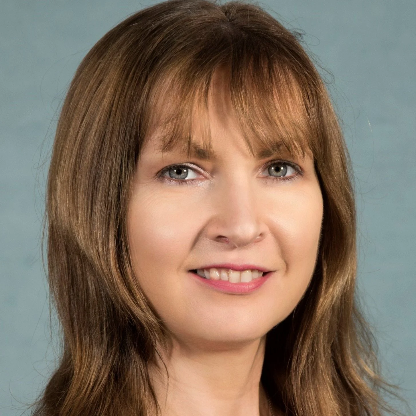 A close-up portrait of a woman with light brown, wavy hair and blue eyes smiling slightly against a gray background.