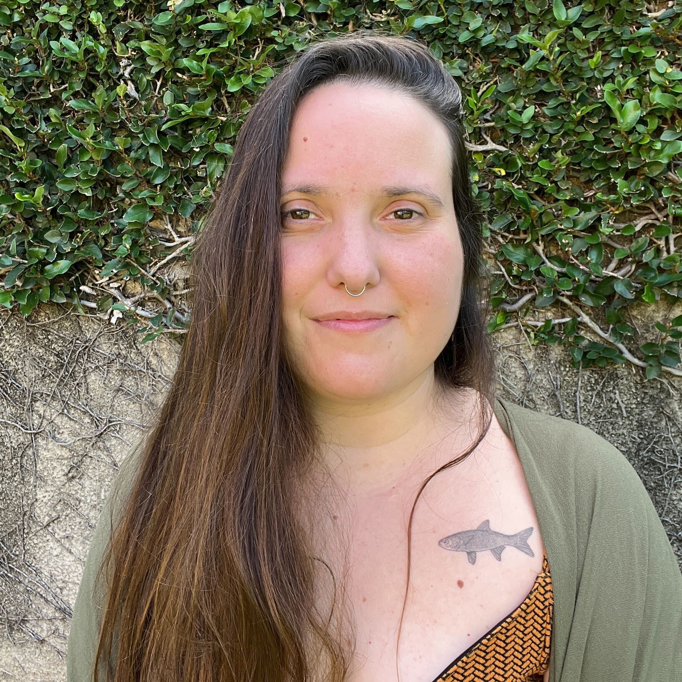 Close-up of a woman with long brown hair, green eyes, and a septum piercing, standing in front of a green leafy hedge. She has a tattoo of a fish on her upper chest and is wearing a necklace with orange beads and an orange and black patterned top.