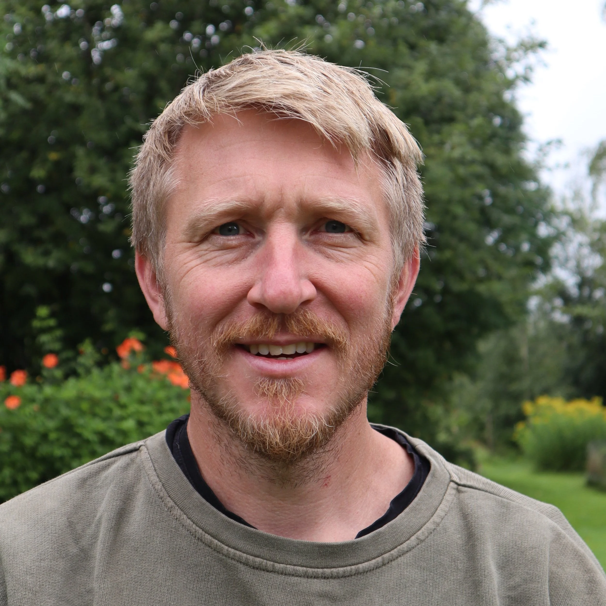 Close-up of a person's face, showing their forehead, eyes, nose, and mouth, with short blonde hair and a beard, outdoors with green trees in the background.