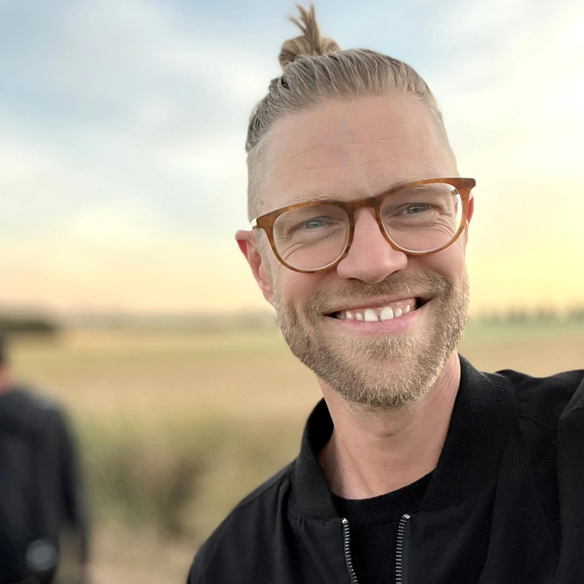 Close-up of a smiling man with blonde hair tied up in a small ponytail, wearing glasses and a black jacket, outdoors on a cloudy day.