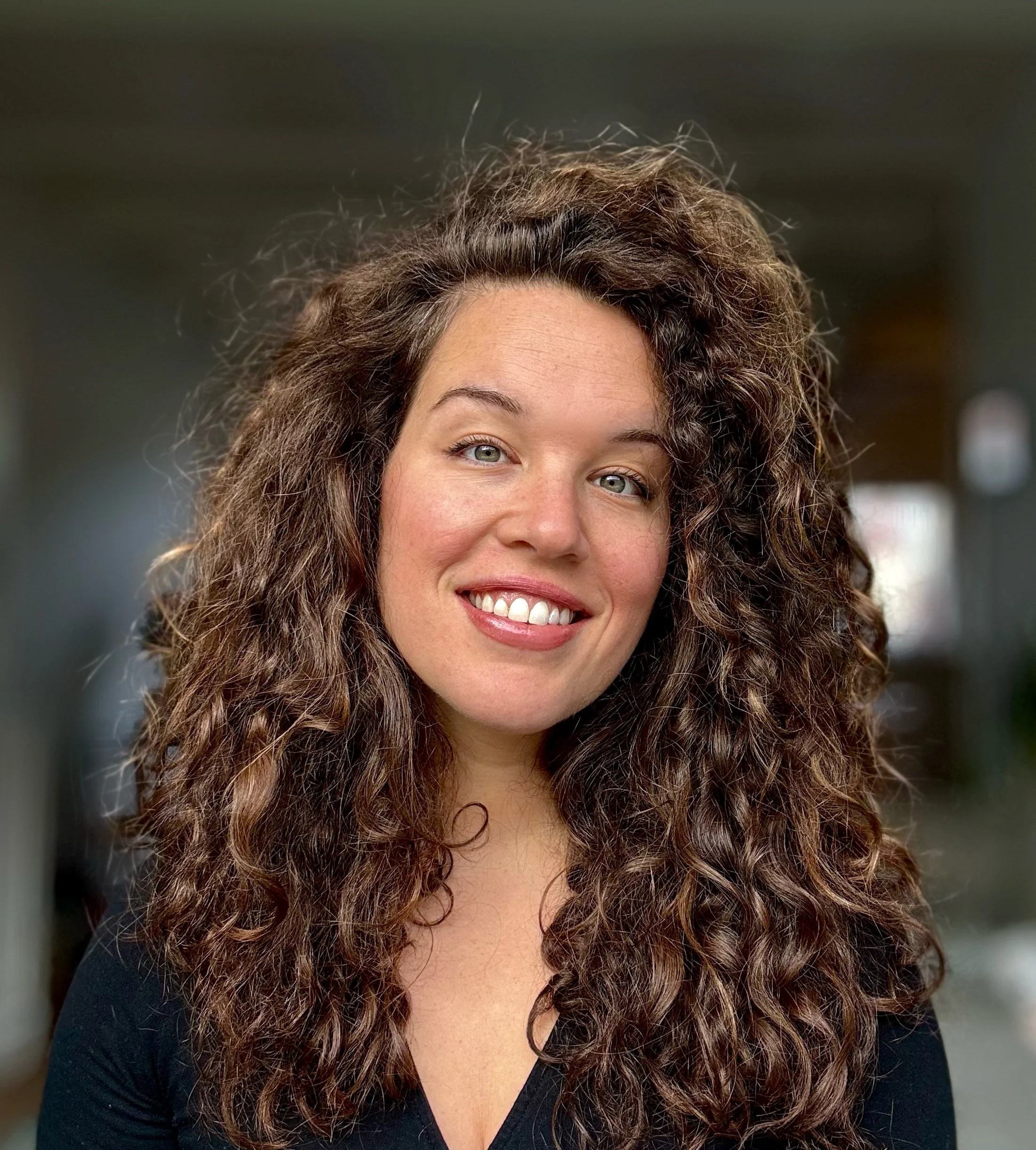 Close-up of a woman with curly brown hair, light skin, and blue eyes, smiling in an indoor setting.