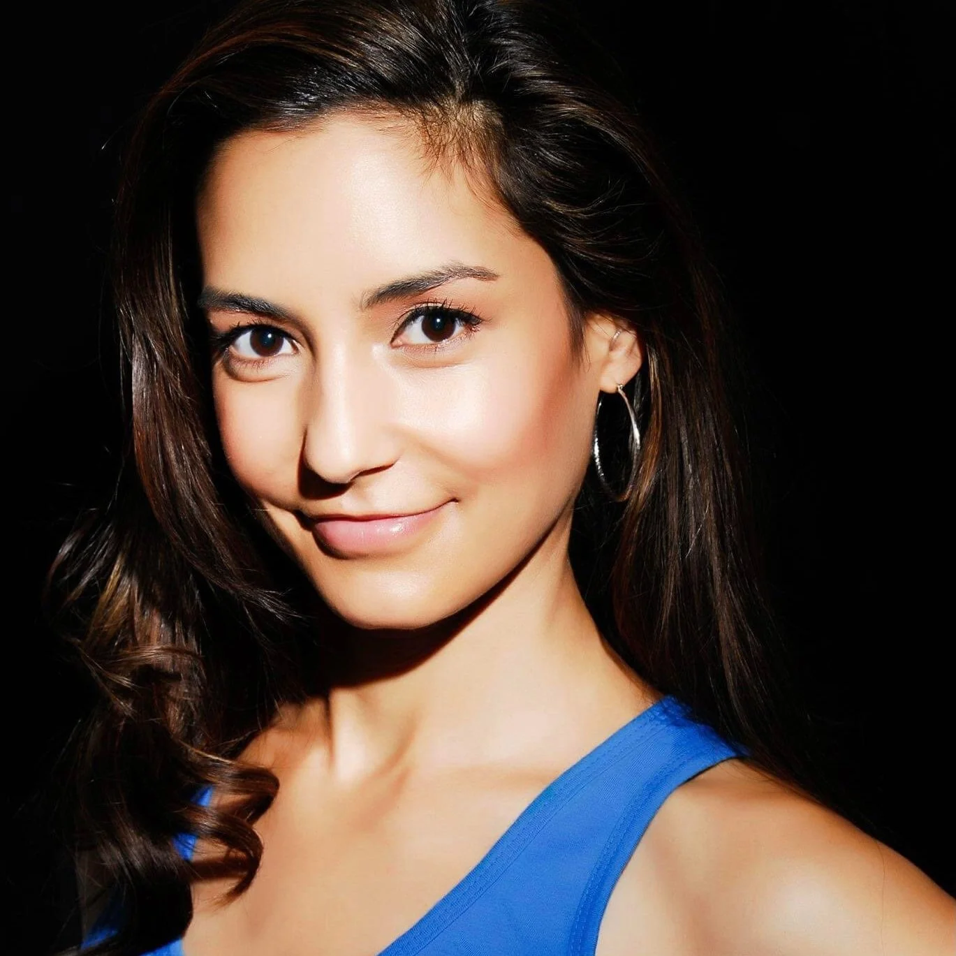 Close-up of a smiling woman with long brown hair, brown eyes, wearing silver hoop earrings and a blue top, against a black background.