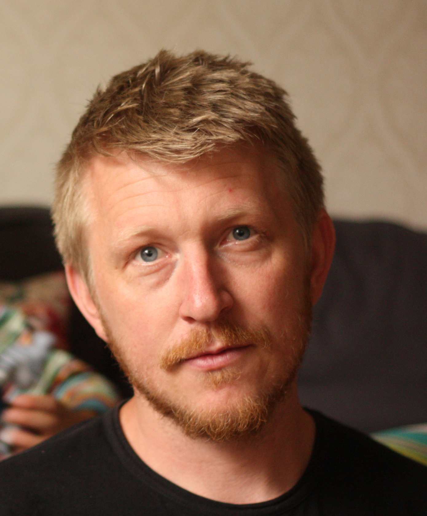 Close-up of a man with blond hair, blue eyes, and a beard, wearing a black shirt, in an indoor setting.