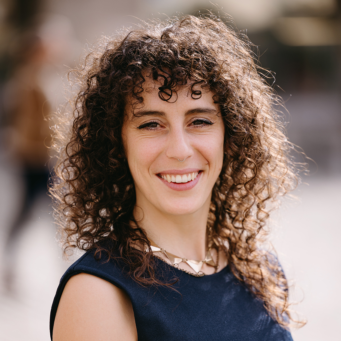 A smiling woman with curly brown hair, wearing a navy sleeveless top and a gold necklace, outdoors on a sunny day.