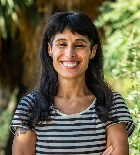 A woman with black hair and a striped t-shirt smiling outdoors with greenery in the background.
