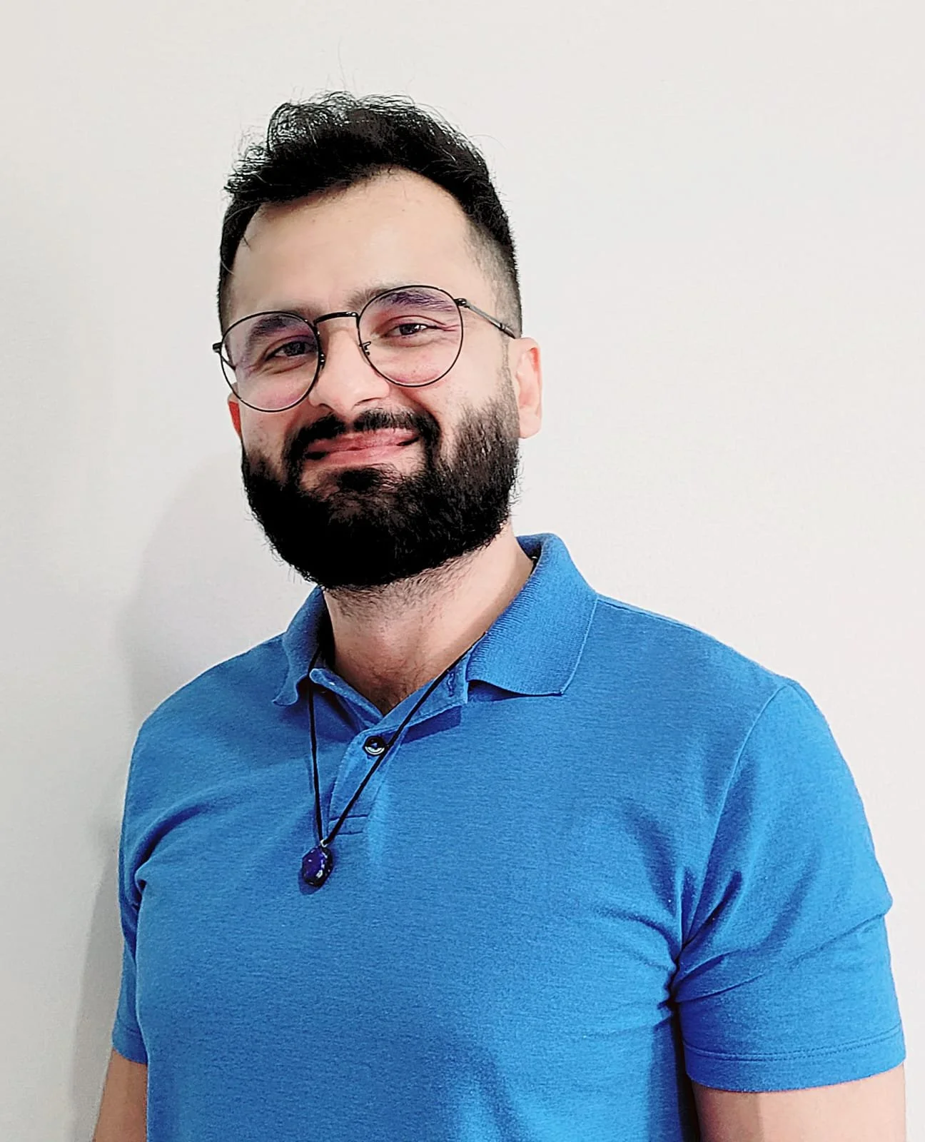 Portrait of a smiling man with dark hair, beard, wearing glasses, a blue polo shirt, and a necklace with a purple pendant, standing against a plain light-colored wall.