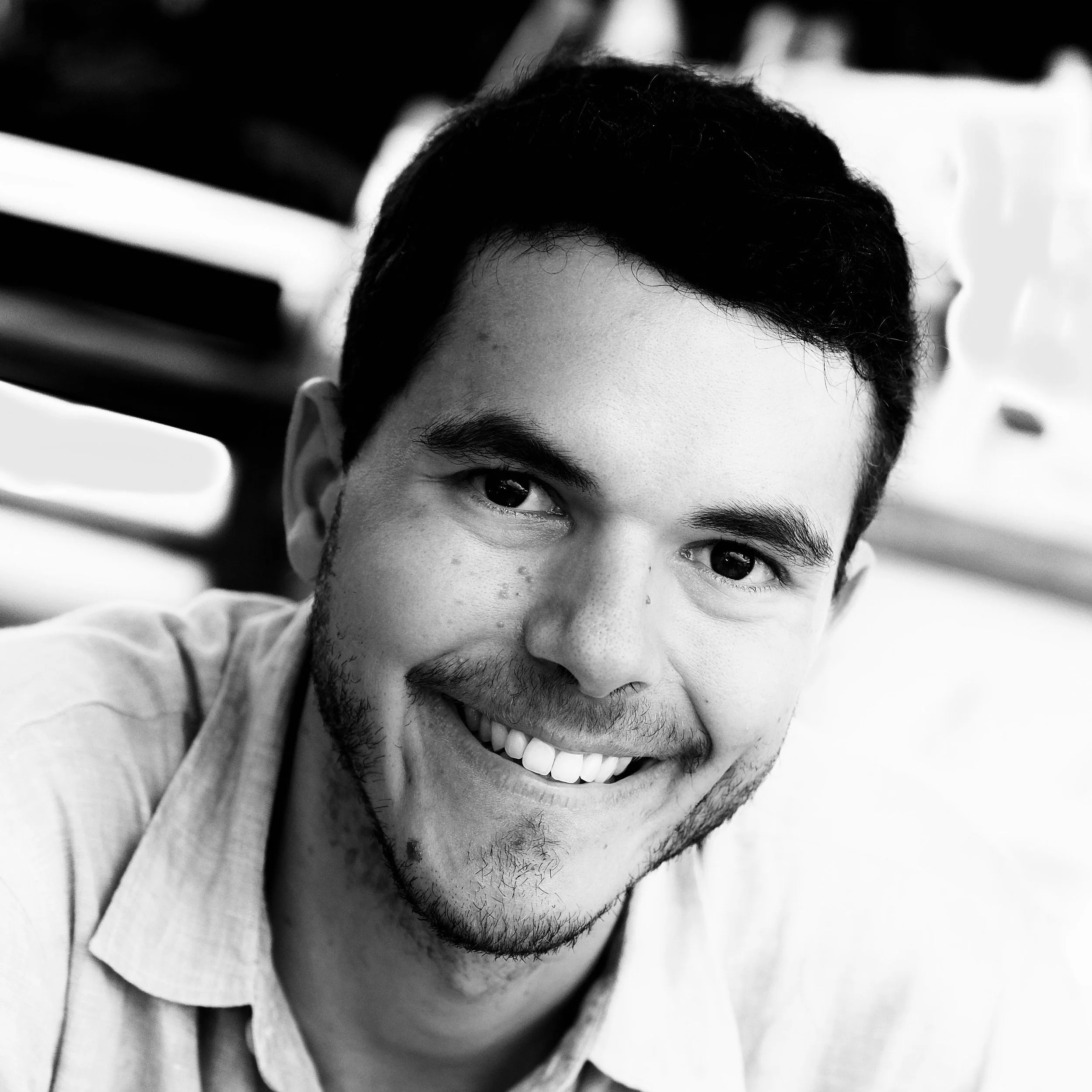 A black and white close-up photo of a young man smiling, showing his teeth and facial hair.