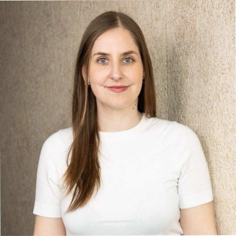 Girl with long brown hair, wearing a white t-shirt, standing against a beige wall, smiling softly.